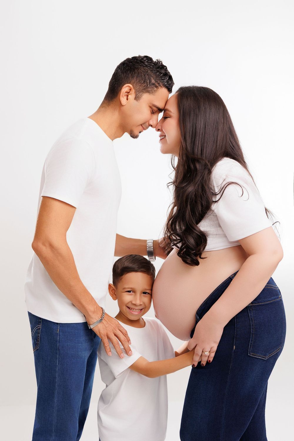 A pregnant woman and man touch foreheads while their young son hugs her baby bump in a family maternity photo.