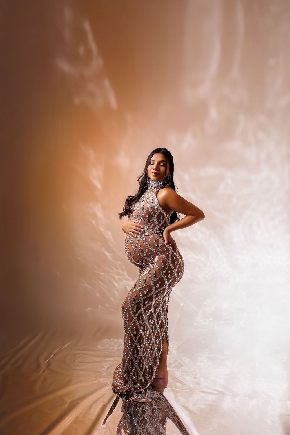 Pregnant woman in elegant sequined gown posing for maternity photo shoot against warm studio backdrop.