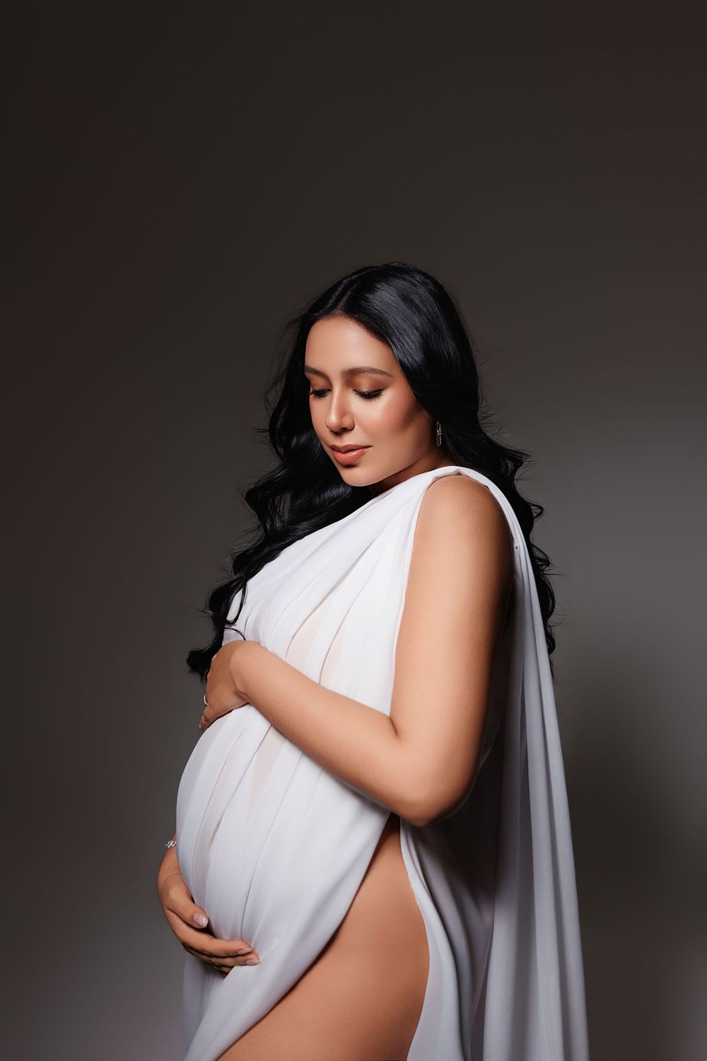 Elegant pregnant woman in white draped fabric gently cradling her baby bump against a dark background.