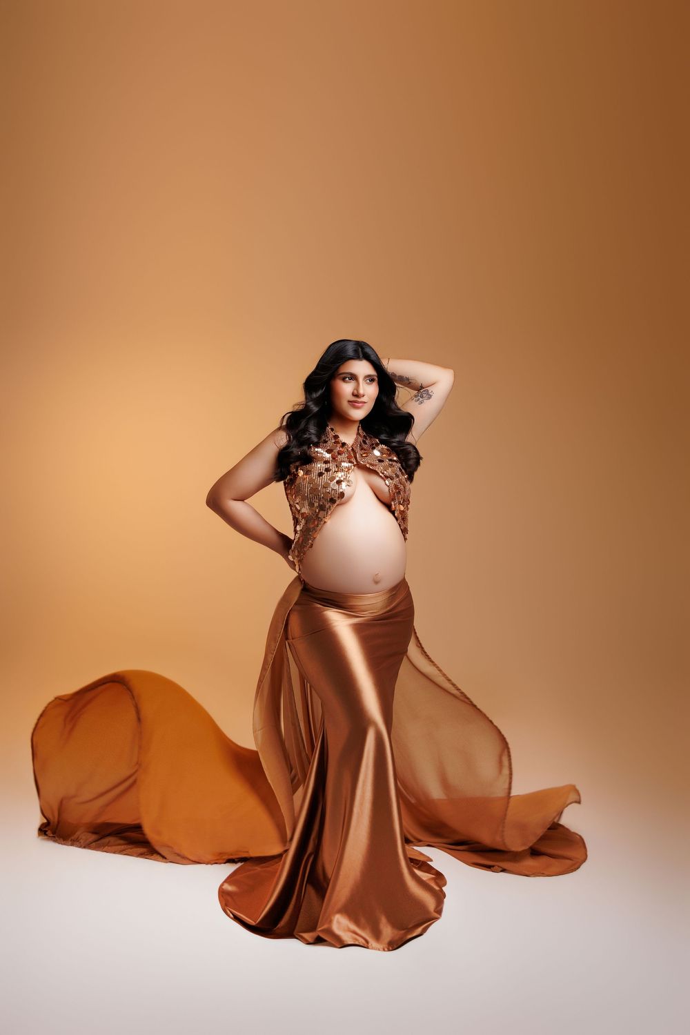 Pregnant woman in elegant gold satin skirt and embellished top poses gracefully against warm beige background.