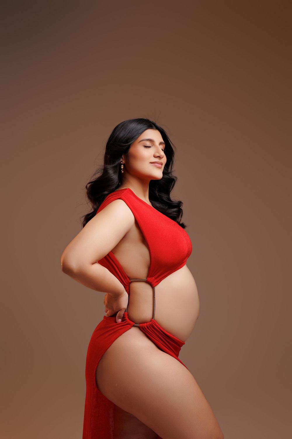 Pregnant woman in red cutout dress posing with hands on hips against brown background.