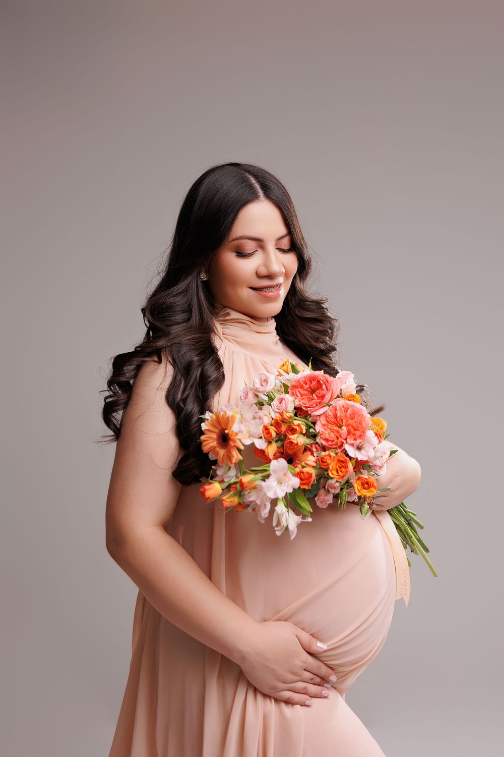 Pregnant woman in peach dress holding orange and pink flower bouquet, smiling with eyes closed in studio.