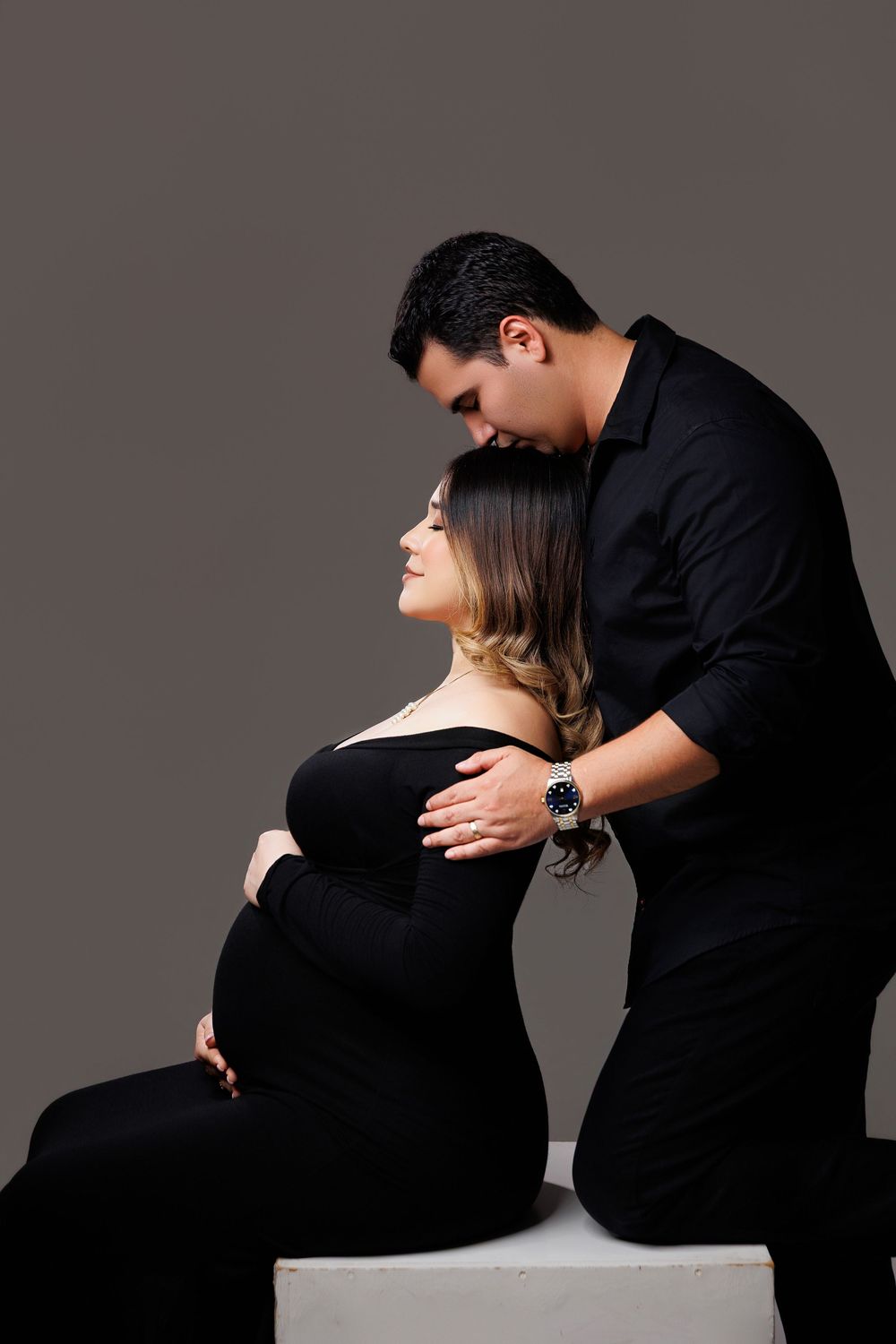 Couple in black maternity photoshoot, man kissing pregnant woman's head while she sits on white cube.