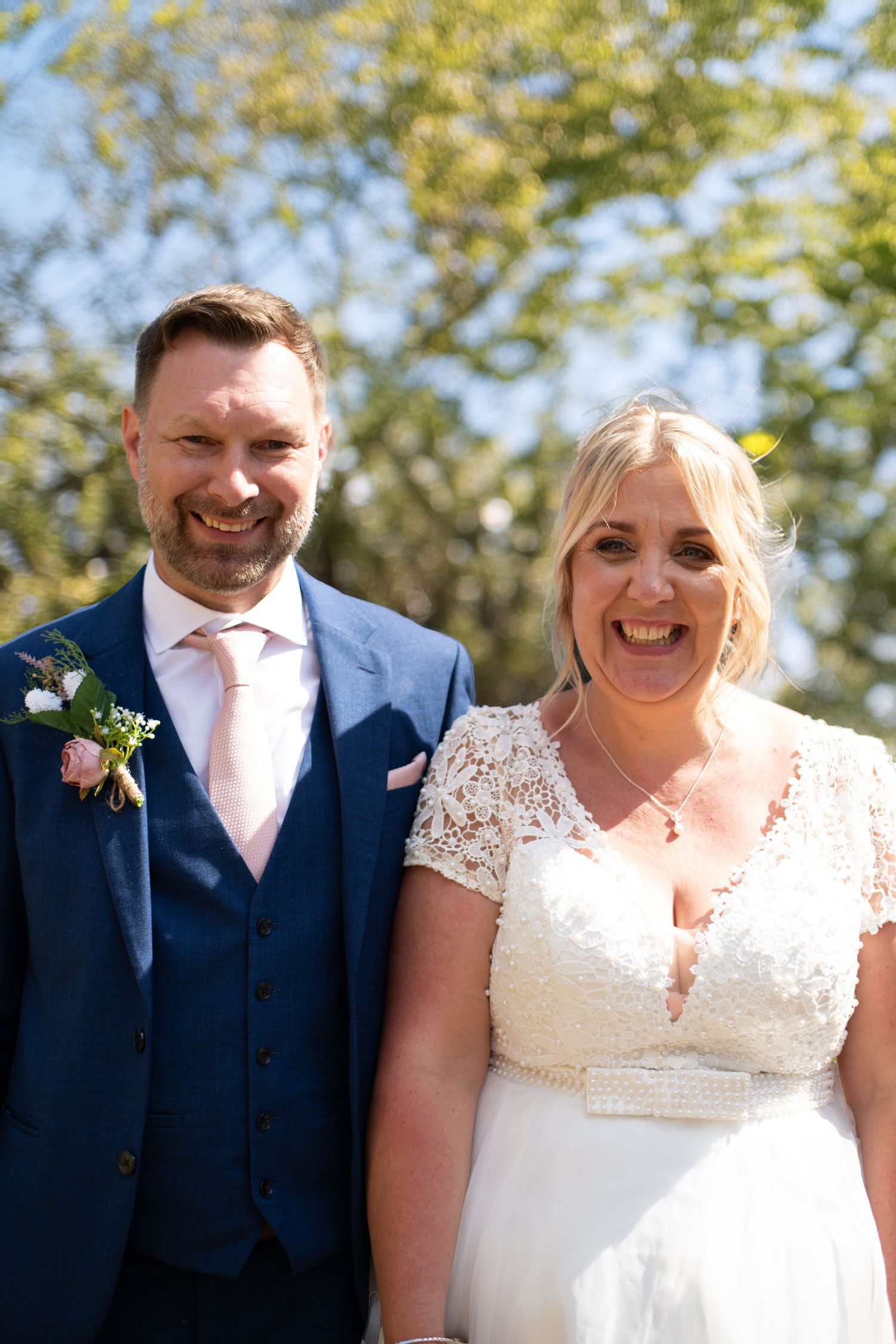 A smiling bride in lace wedding dress and groom in navy blue suit with pink tie pose outdoors on their wedding day.