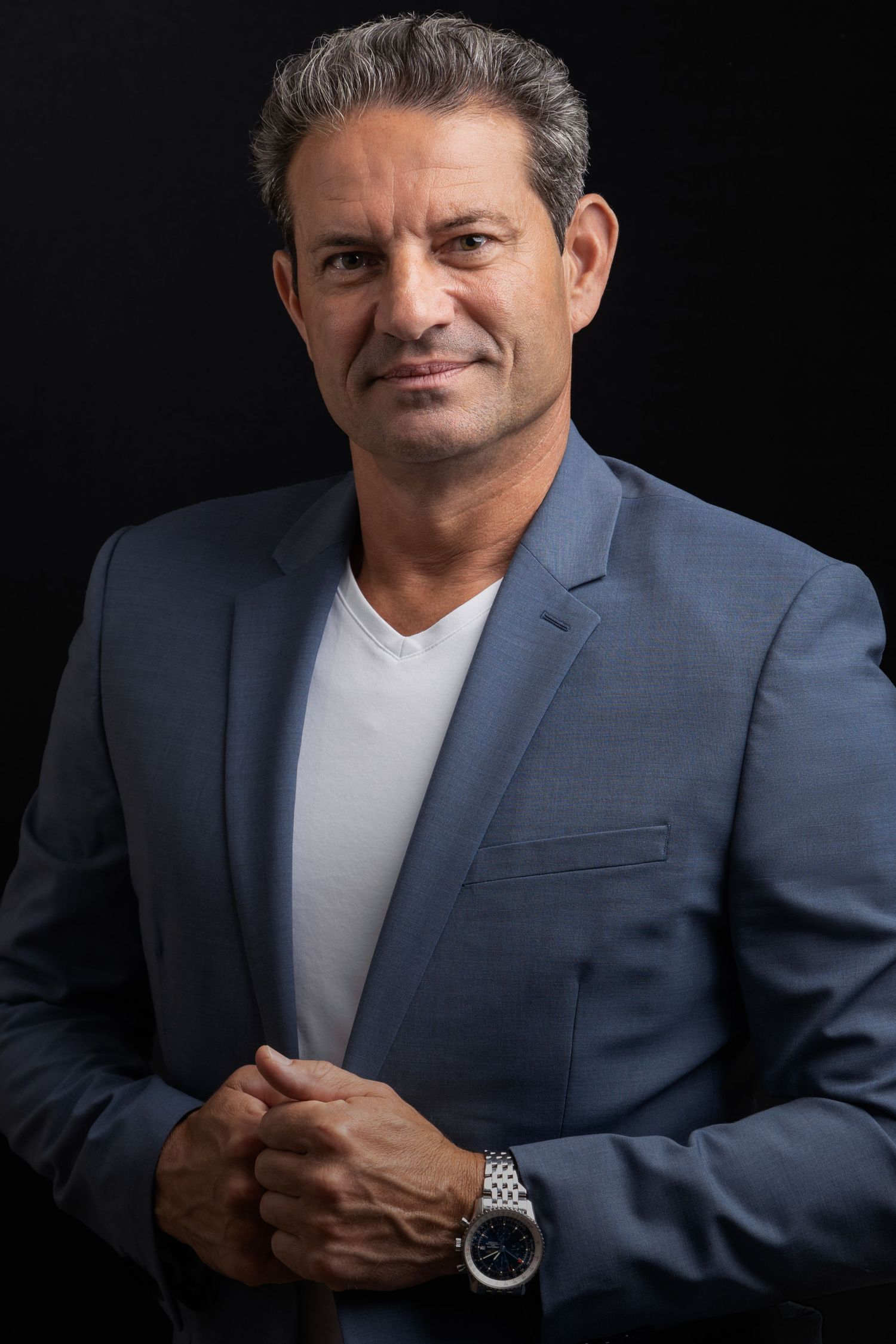 Middle-aged man in blue blazer and white t-shirt, wearing a watch, posing confidently against dark background.