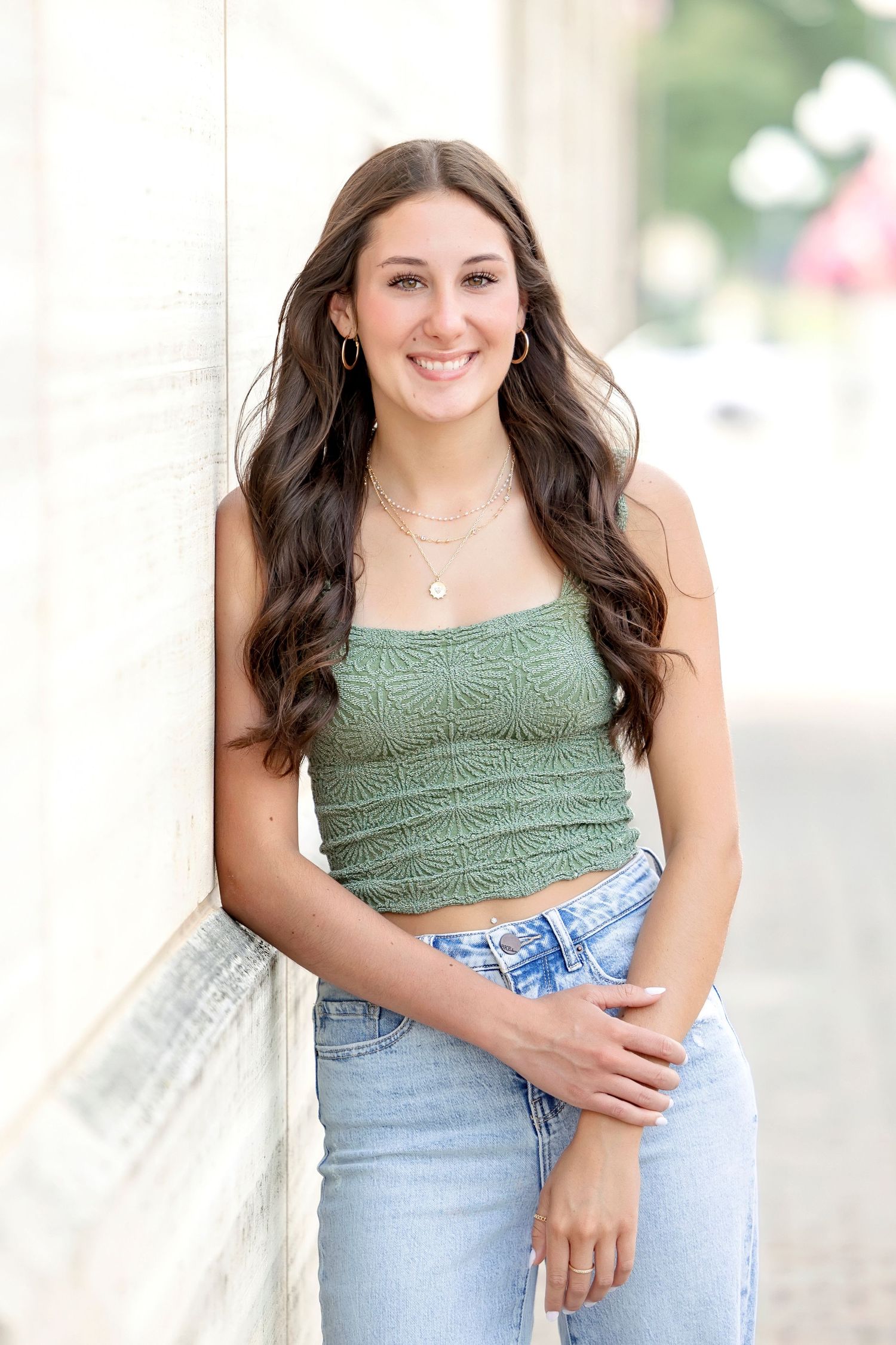 Smiling teenage girl in green crop top and jeans leans against white wall for senior portrait photo.