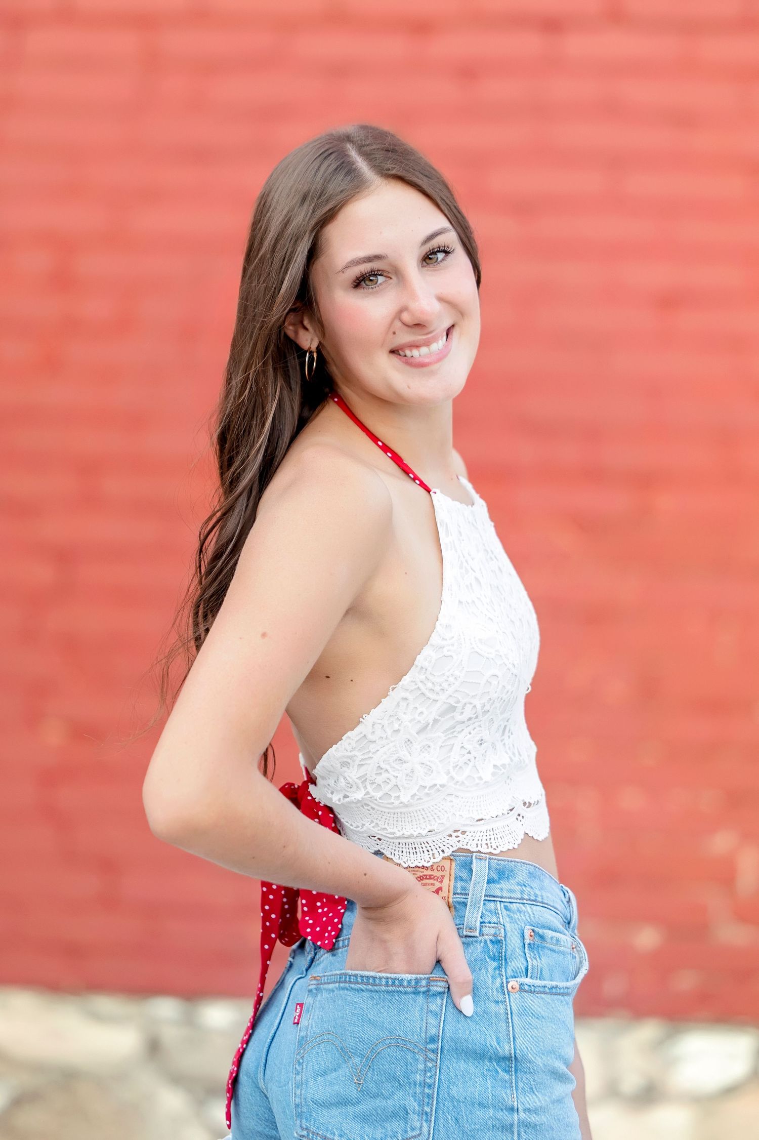 Young woman in white lace crop top and denim shorts smiling against a red brick wall.