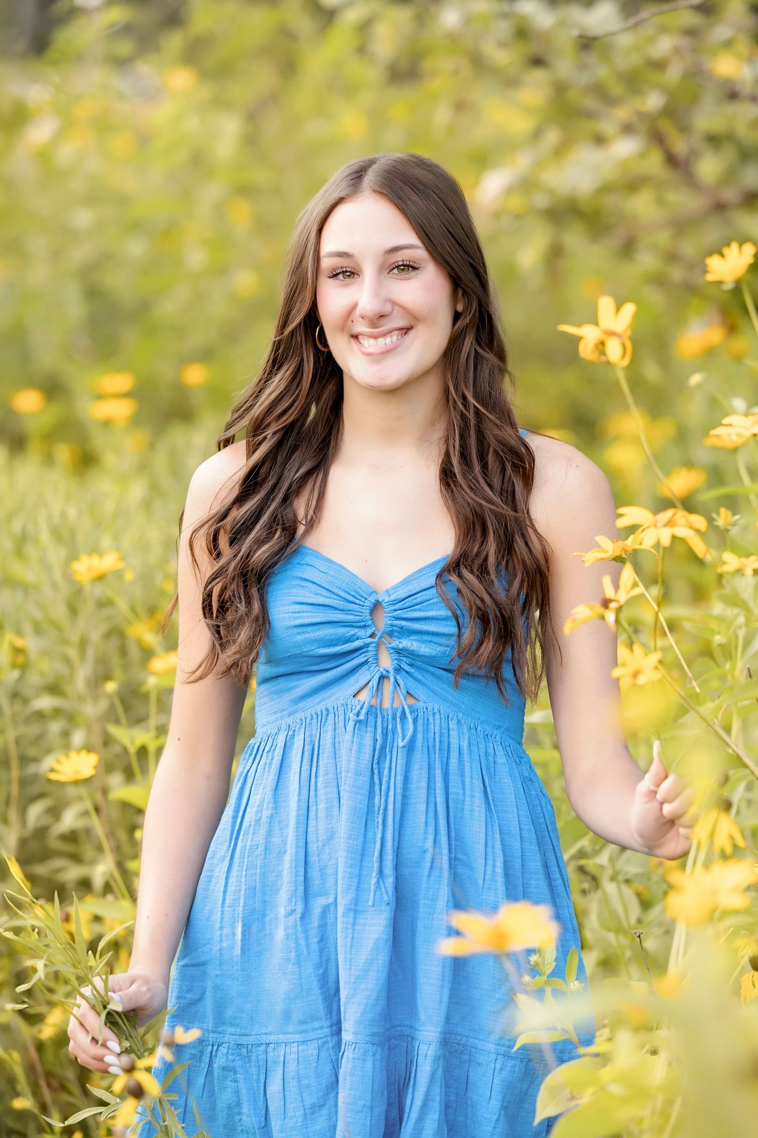 Smiling young woman in blue sundress standing among vibrant yellow wildflowers in a garden.