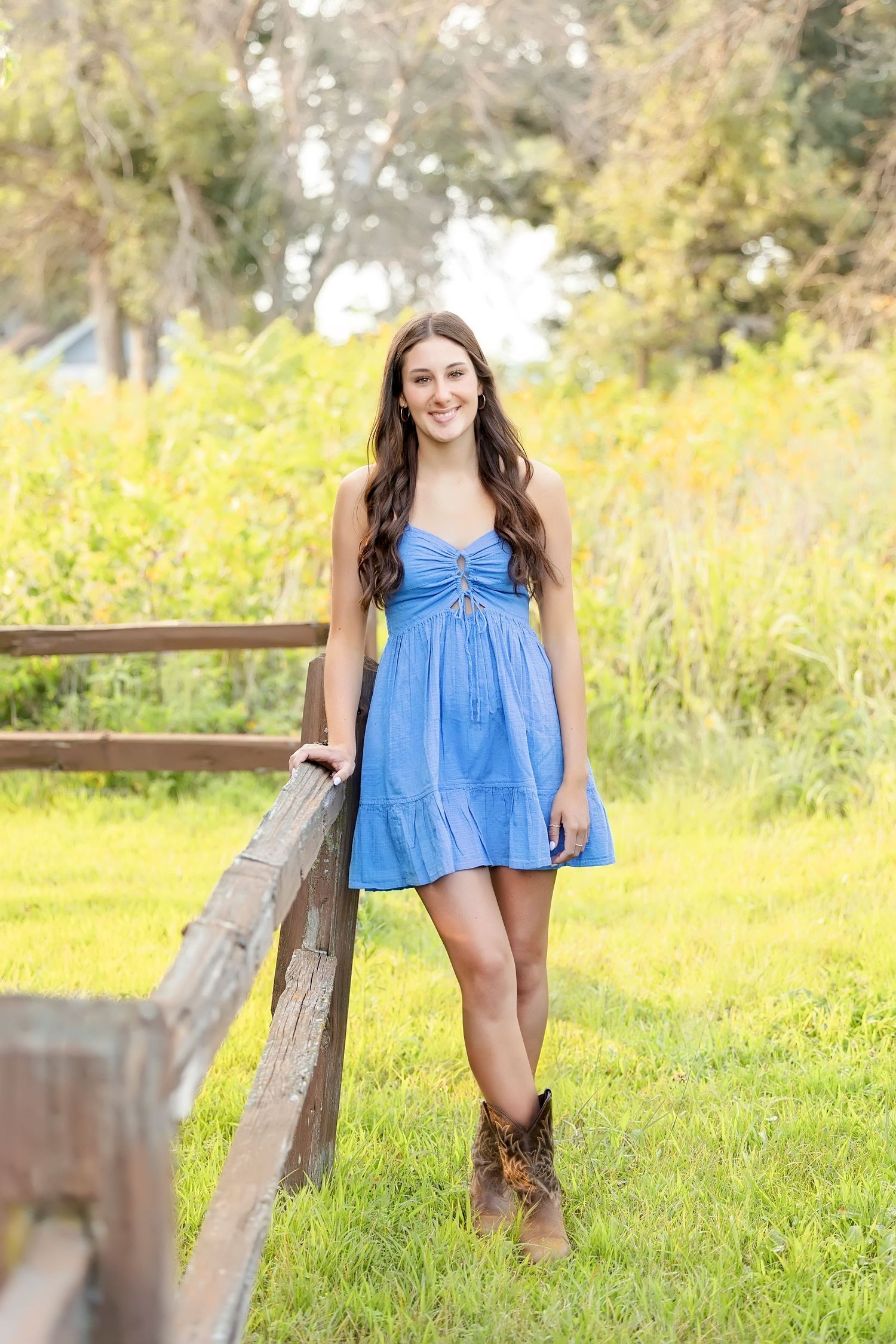 Young woman in blue sundress and cowboy boots leaning on a wooden fence in a sunny field.