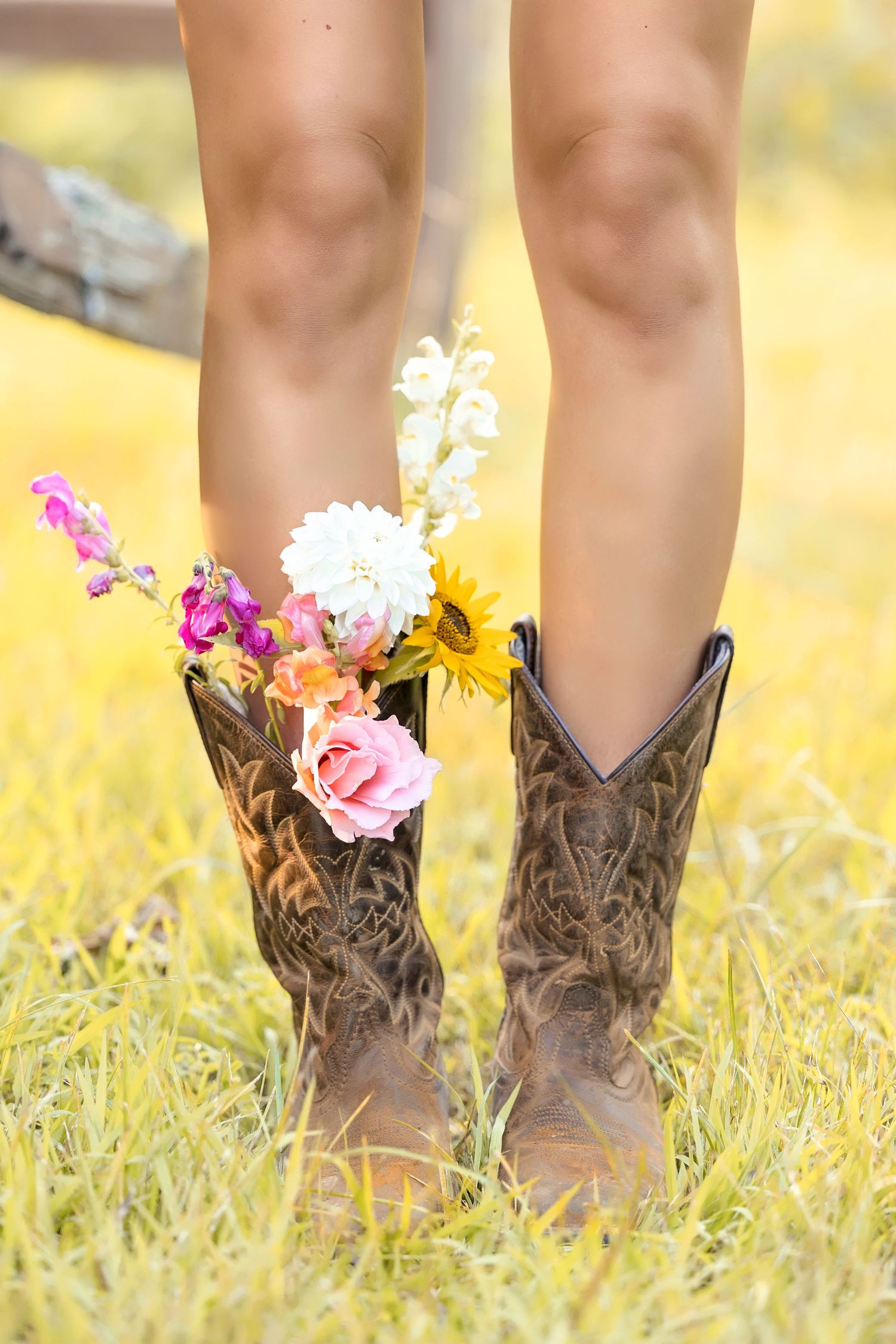 Person wearing brown cowboy boots filled with colorful wildflowers standing in a sunny yellow grass field.