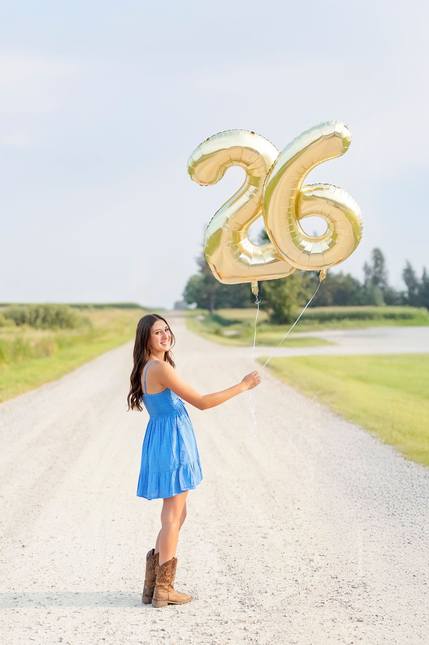 Young woman in blue dress and cowboy boots holds gold number 26 balloons on a rural gravel road.