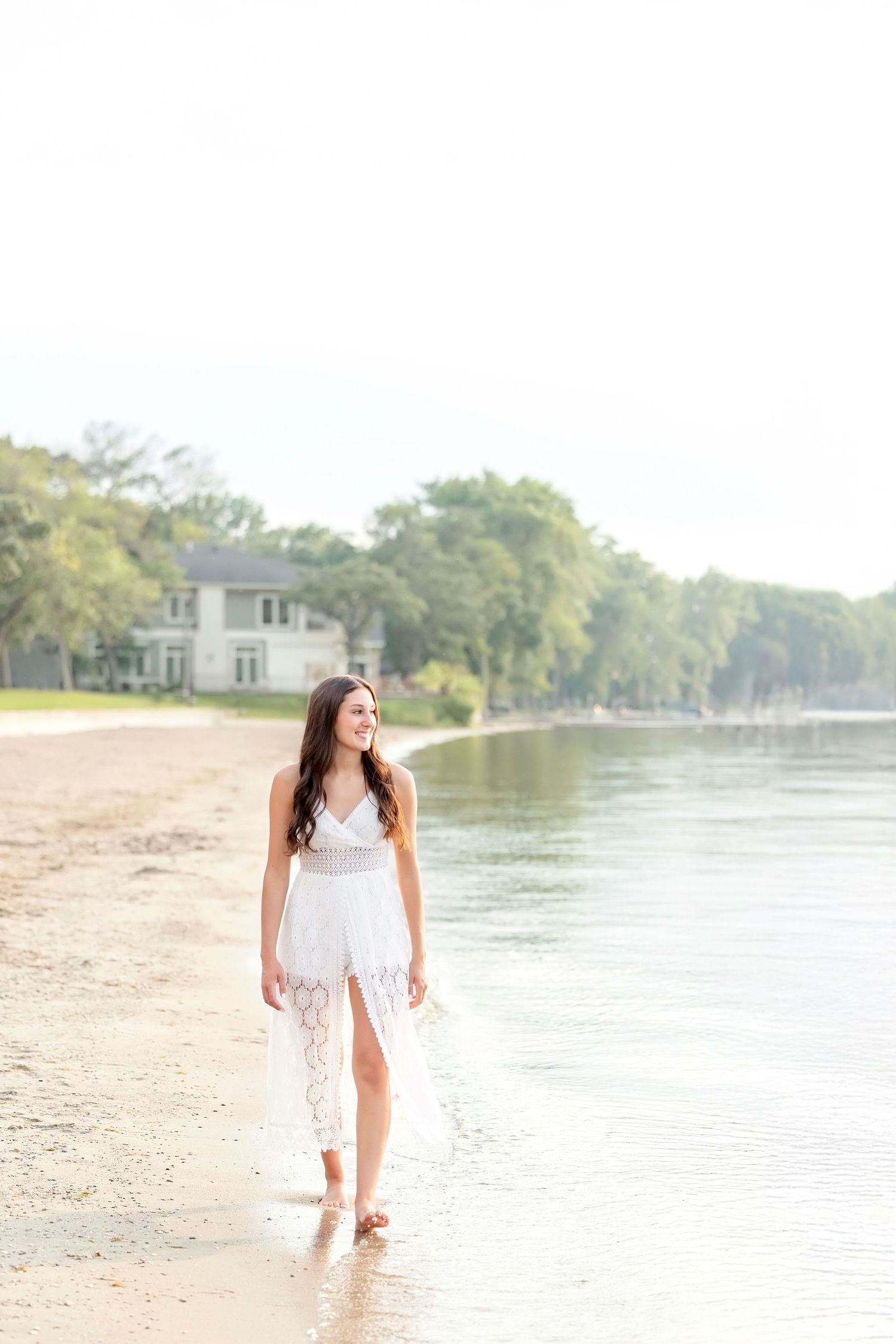 Brunette young woman in white lace dress walking barefoot along a serene lakeside beach shoreline.