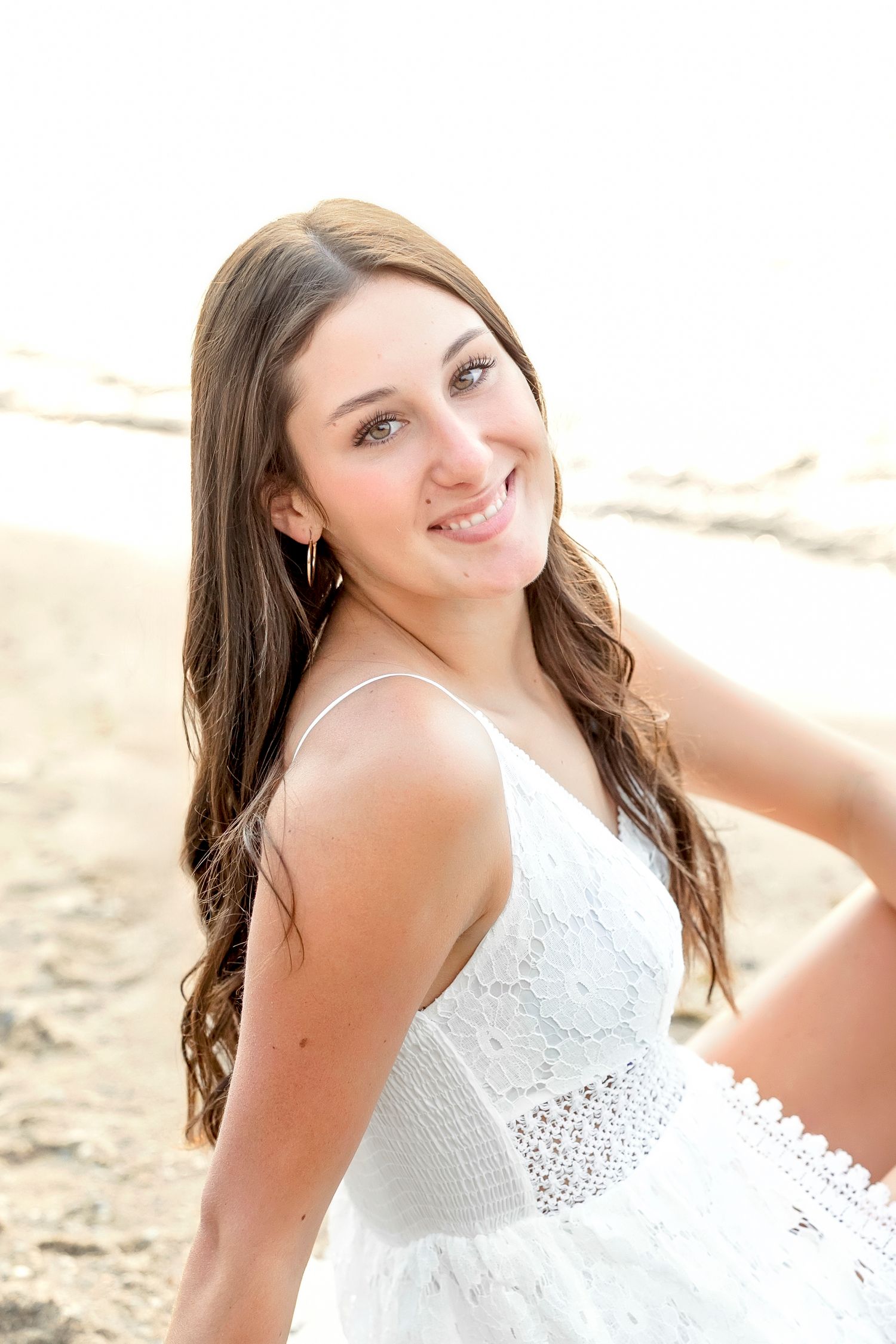 Young woman with long brown hair smiling in a white lace dress at the beach.