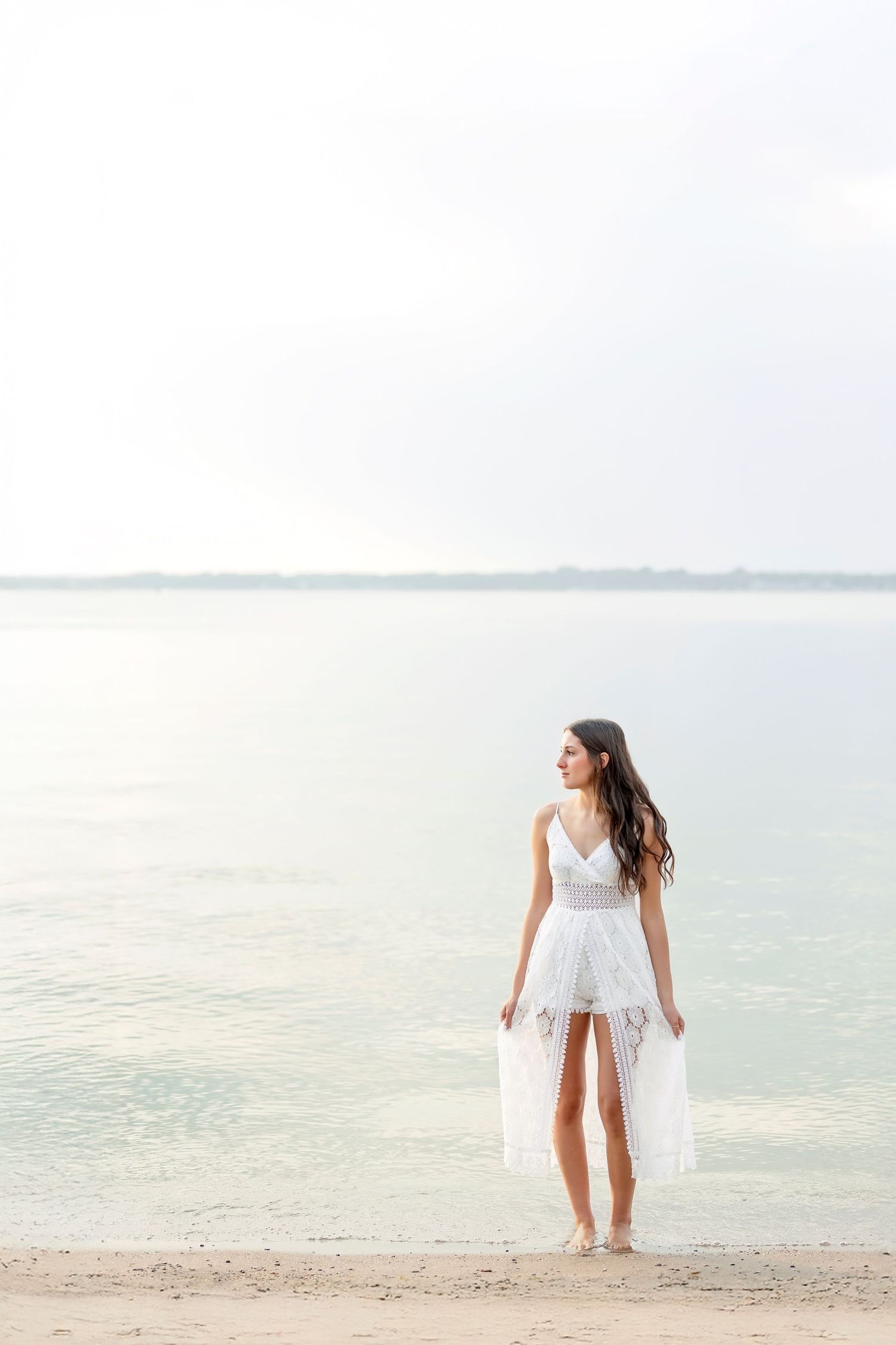 Woman in white lace dress standing on sandy beach by calm water, looking sideways in soft natural light.