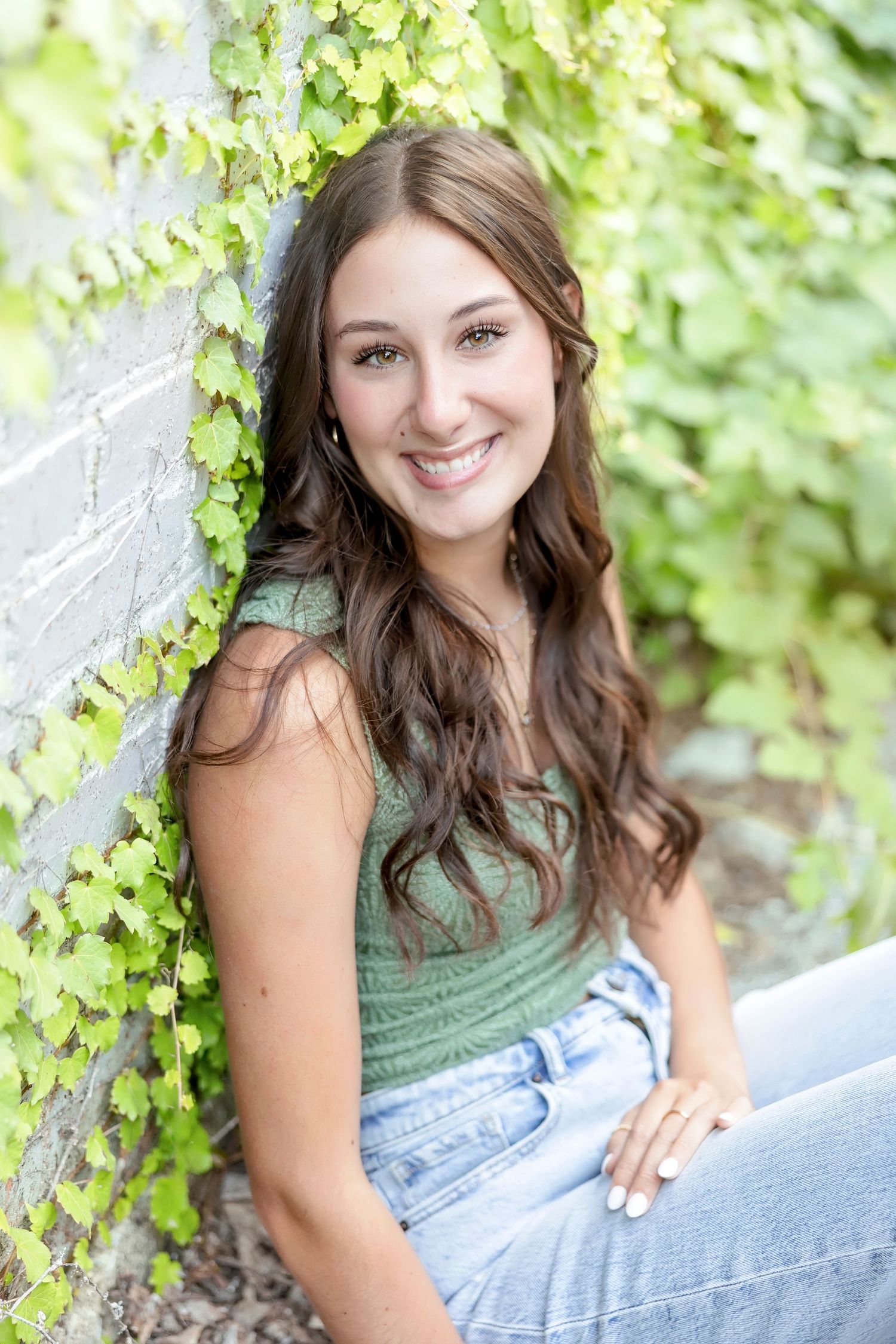Young woman with long brown hair smiling in green ivy-covered wall setting, wearing green top and jeans.
