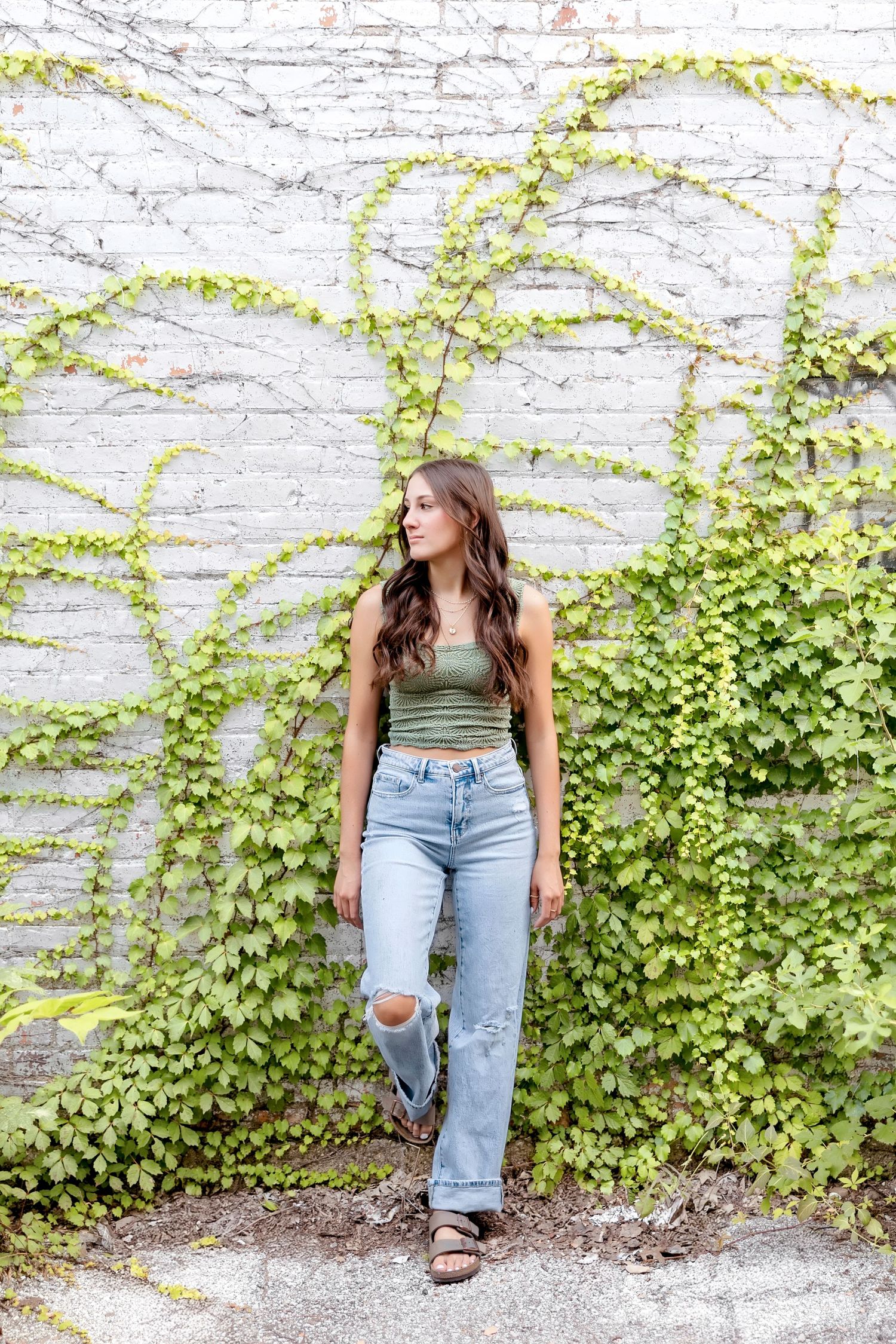 Young woman in green tank top and ripped jeans leaning against ivy-covered white brick wall.