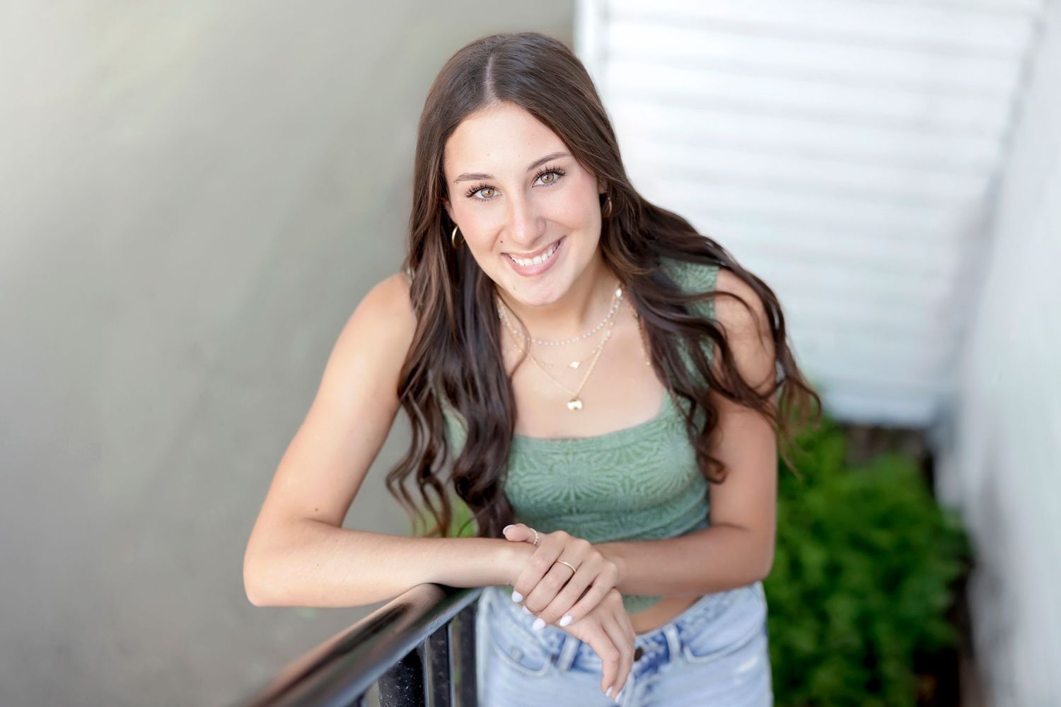 Young woman with long brown hair smiling outdoors, wearing a green crop top and jeans, leaning on a railing.