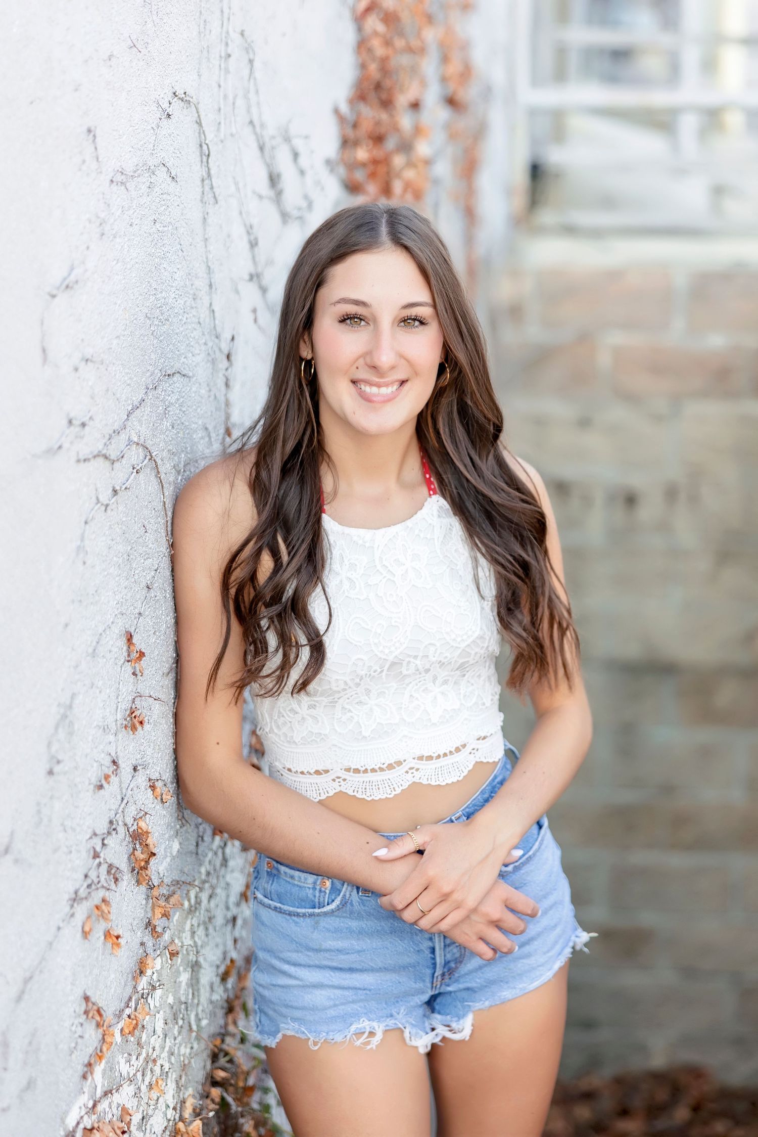 Smiling brunette teen in white lace crop top and denim shorts leaning against a white textured wall.