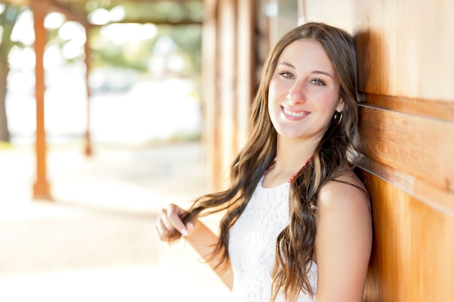 Smiling young woman with long brown hair leaning against a wooden wall outdoors in a white dress.
