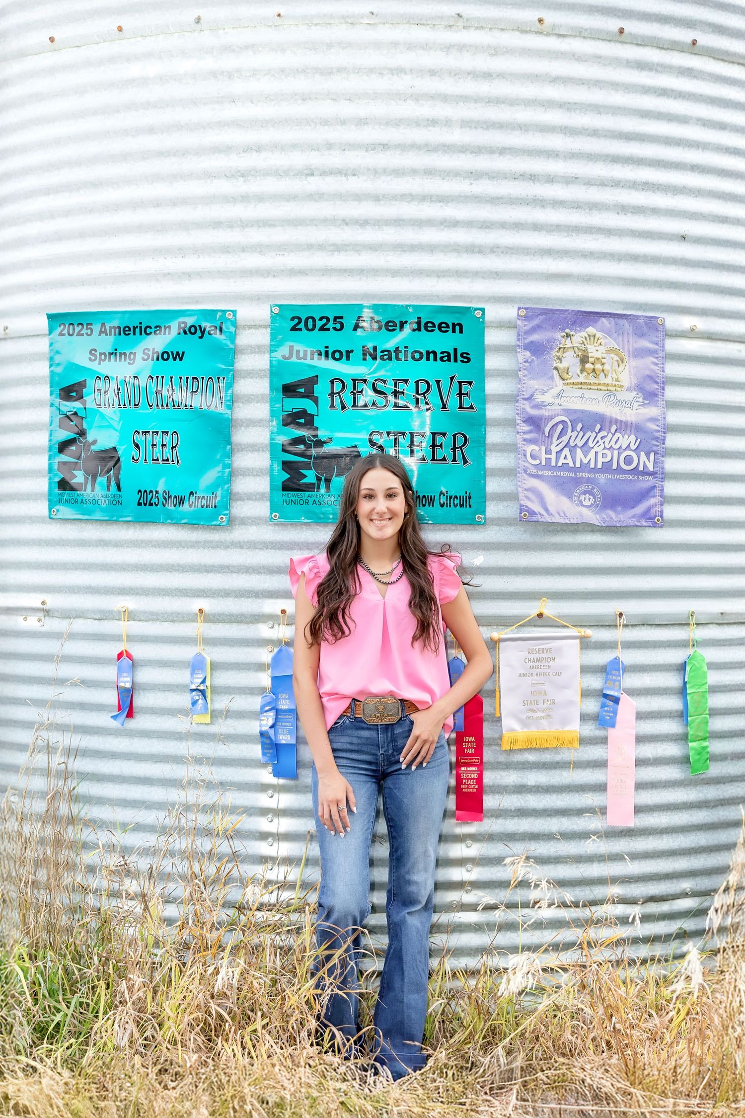 Young woman in pink top and jeans stands proudly before livestock show championship banners on a grain silo.