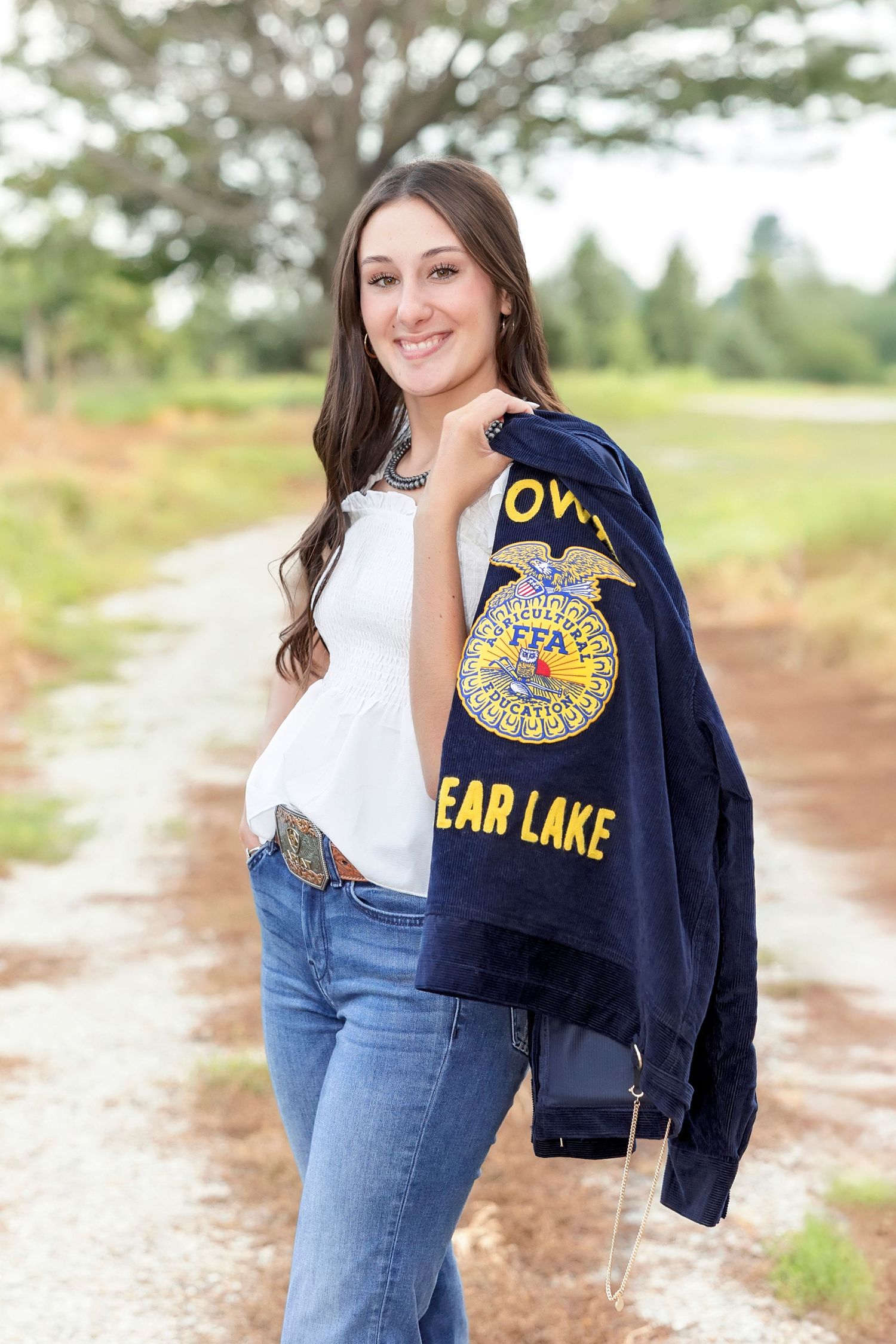 Young woman smiling in FFA jacket over shoulder, jeans, and western belt on a scenic dirt path outdoors.