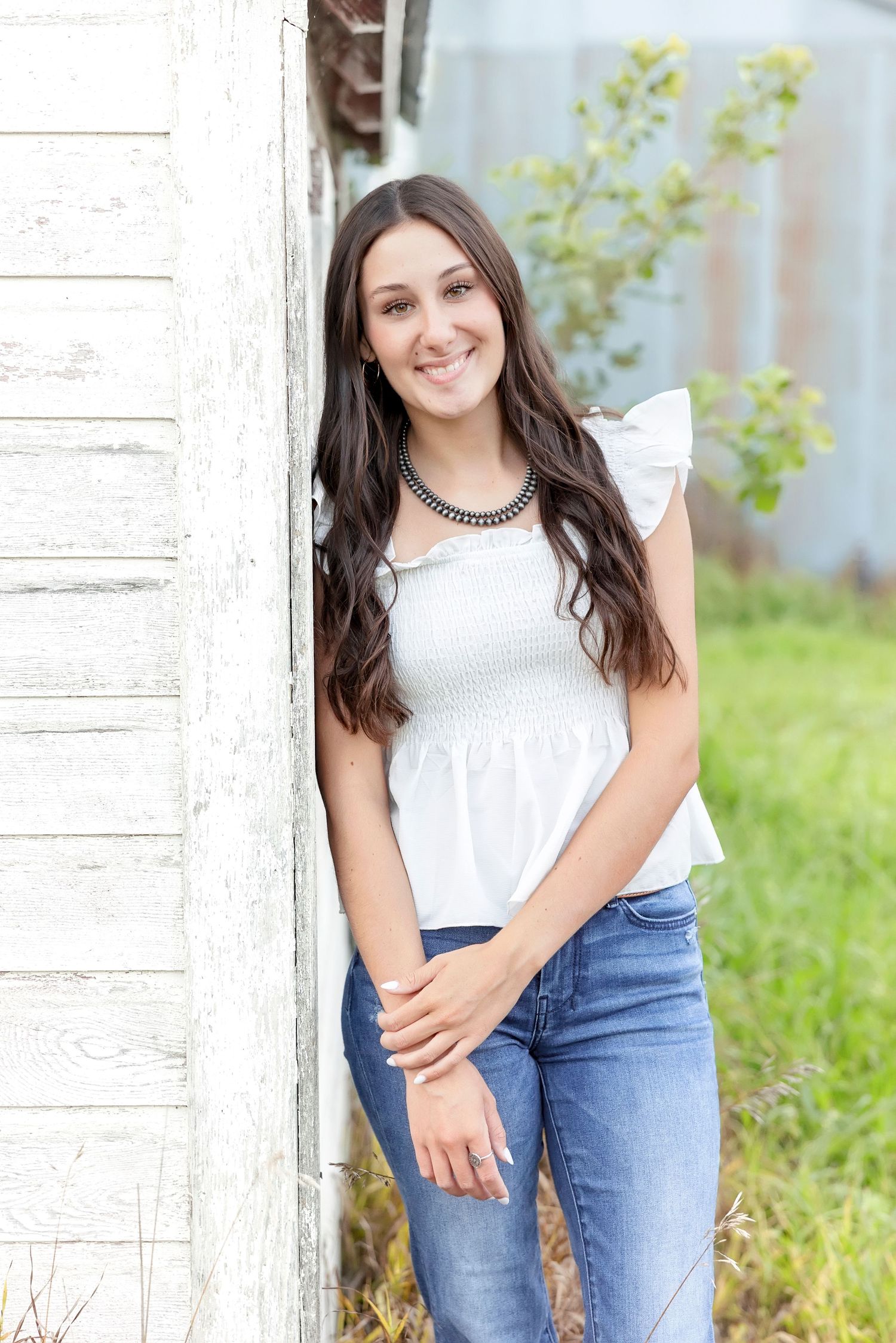 Smiling brunette teen in white ruffled top and jeans posing by rustic white wooden door outdoors.
