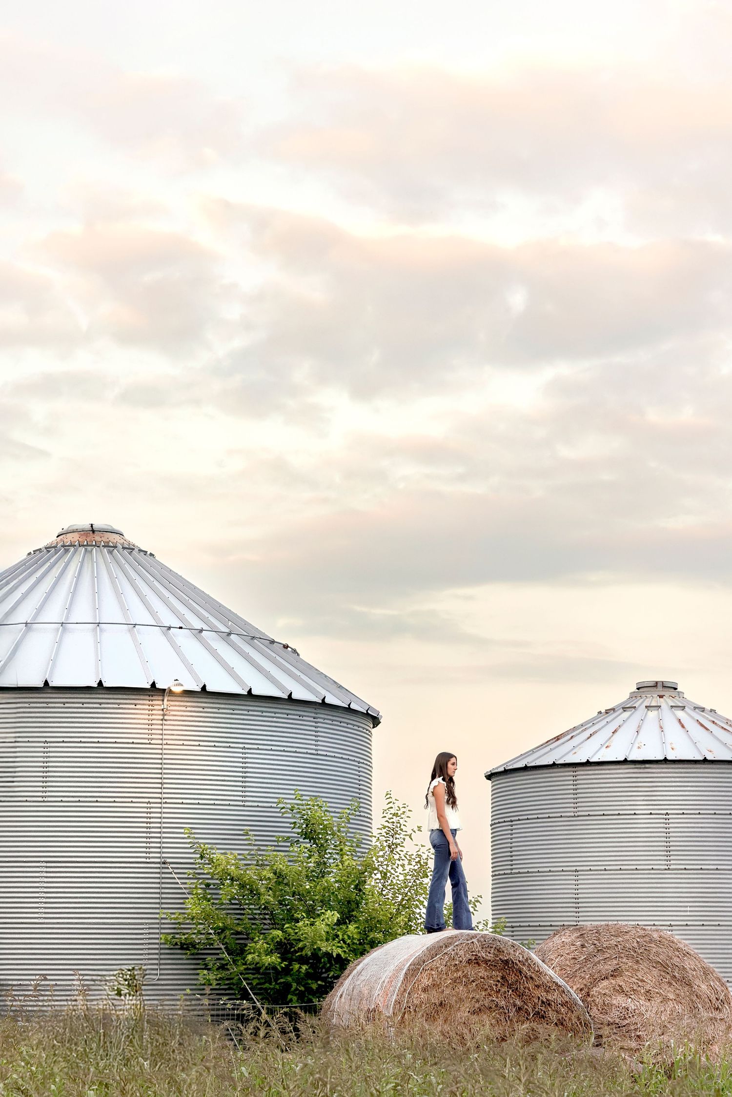 A young woman stands on a hay bale between two large grain silos on a farm under a cloudy sky.