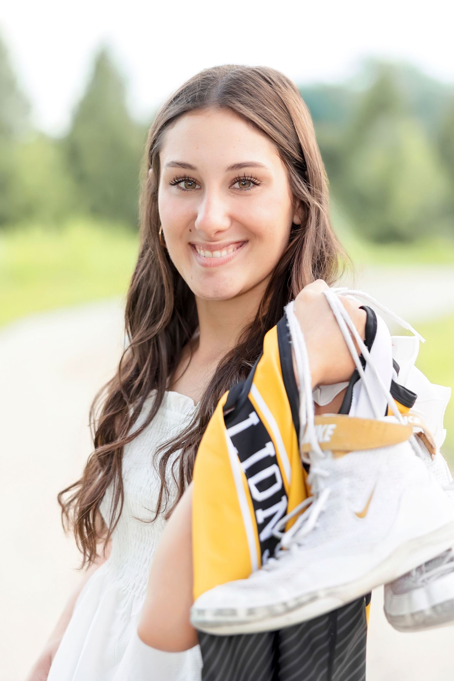 Smiling young woman with long brown hair holding white Nike skates and a yellow scarf outdoors.