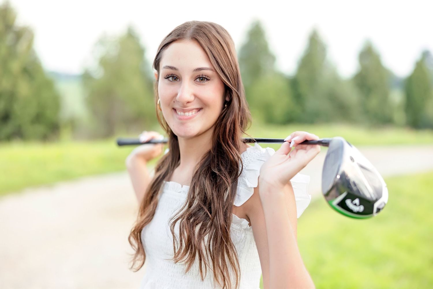 Young woman smiling on golf course, holding a golf club over her shoulder, wearing a white top outdoors.