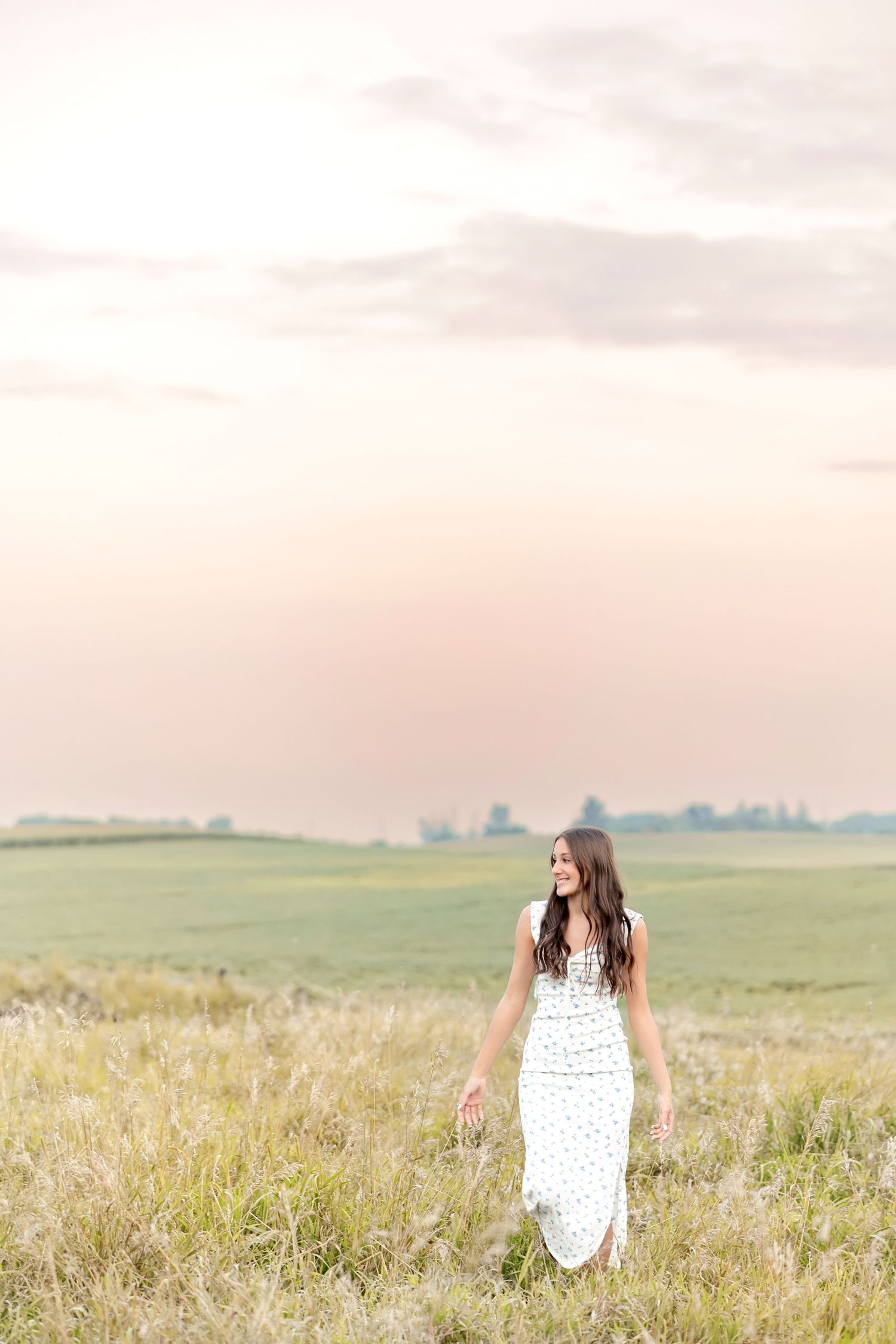Young woman in white maxi dress walking through a green field at sunset.