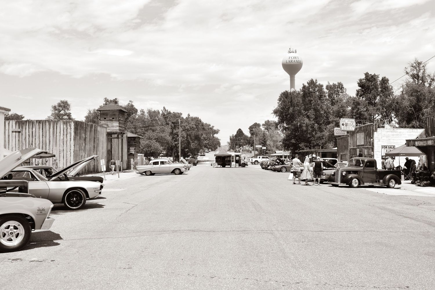 Vintage black and white photo of a small-town main street with classic cars parked along the road and a water tower.