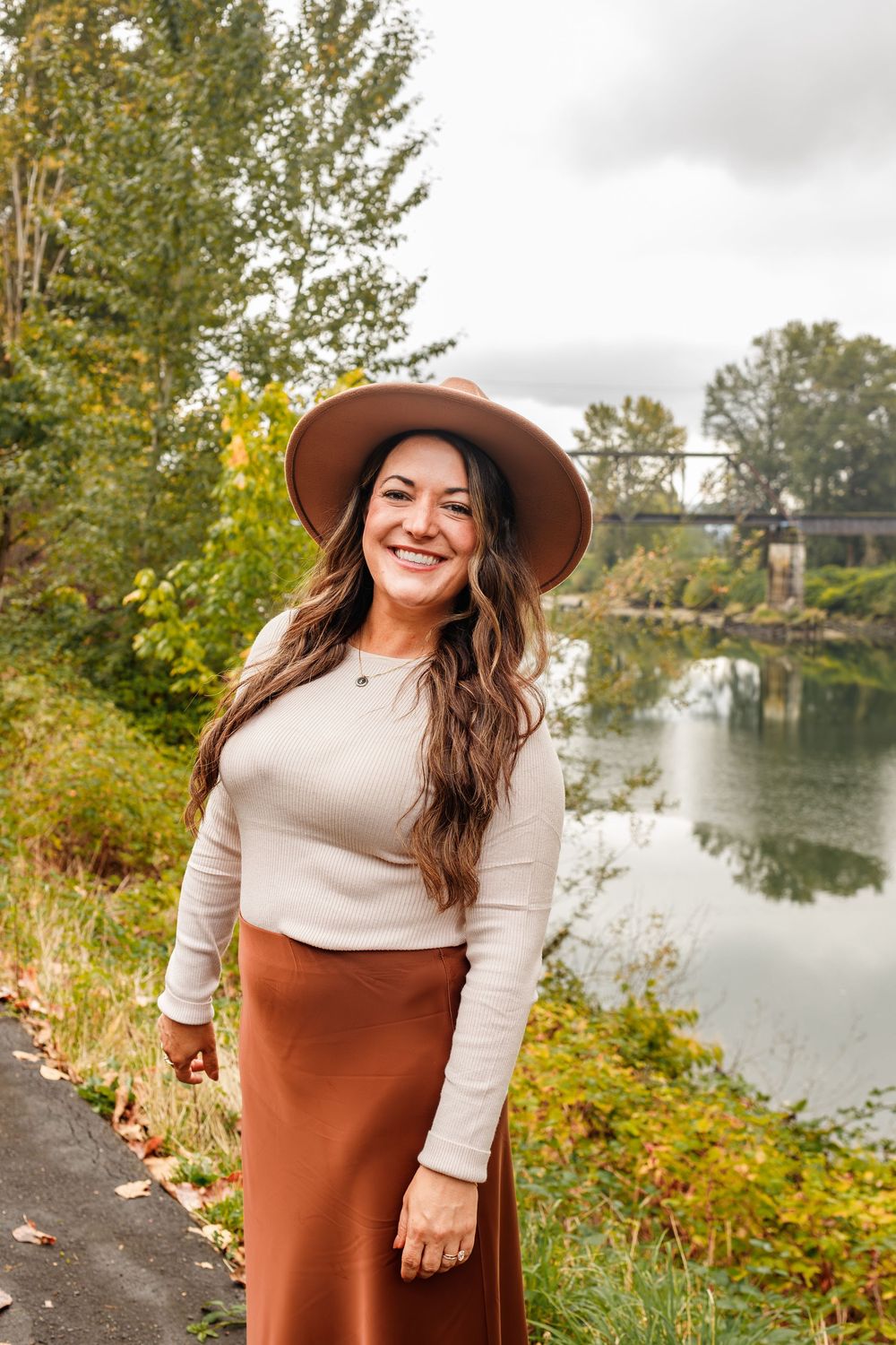 Woman in tan hat and rust skirt smiling by a river with autumn trees and bridge in background.