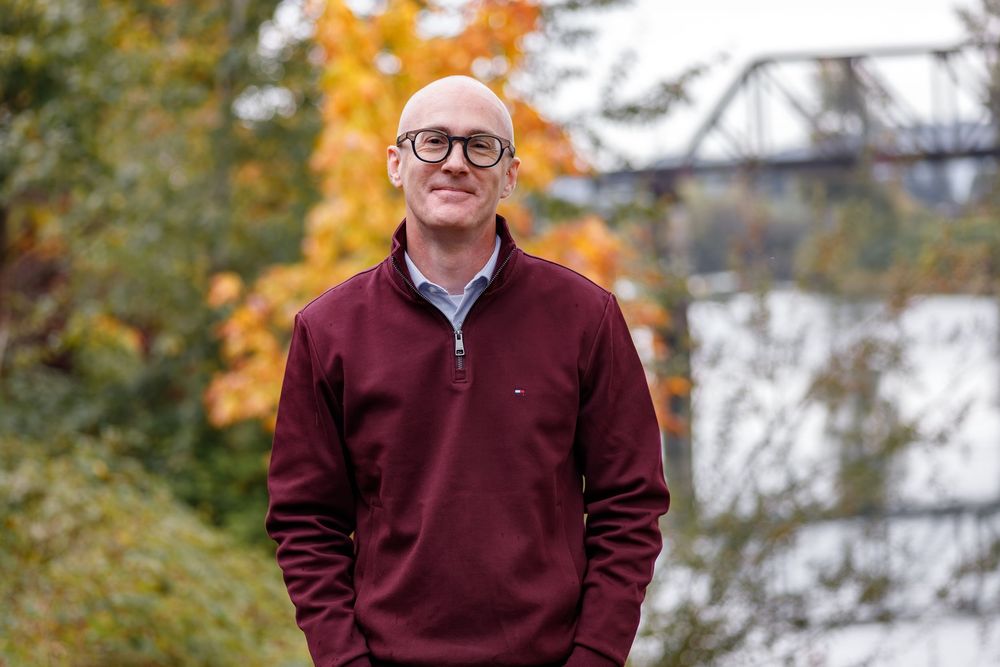 Bald man with glasses wearing a maroon Tommy Hilfiger pullover standing outdoors in autumn foliage near a river.