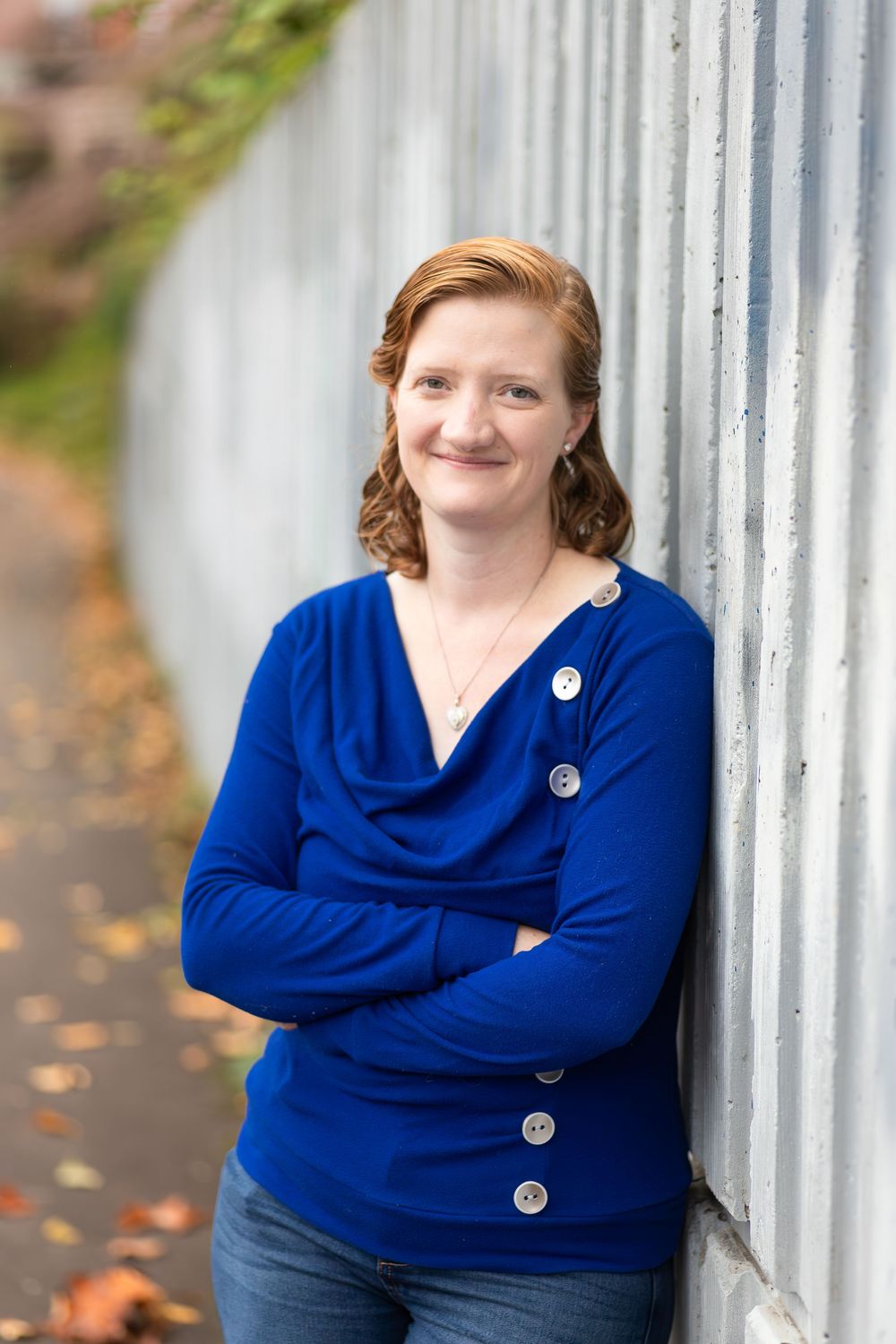 Woman in blue sweater with arms crossed leaning against a wall outdoors in autumn.