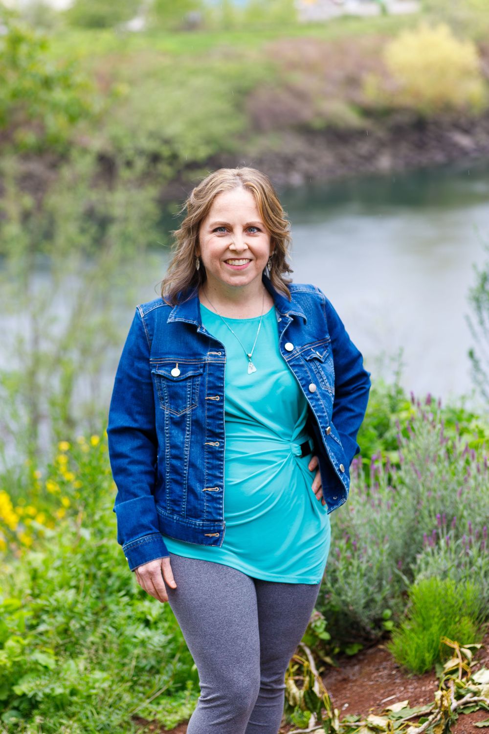 Smiling woman in denim jacket and teal shirt posing outdoors near a river with lush green vegetation.