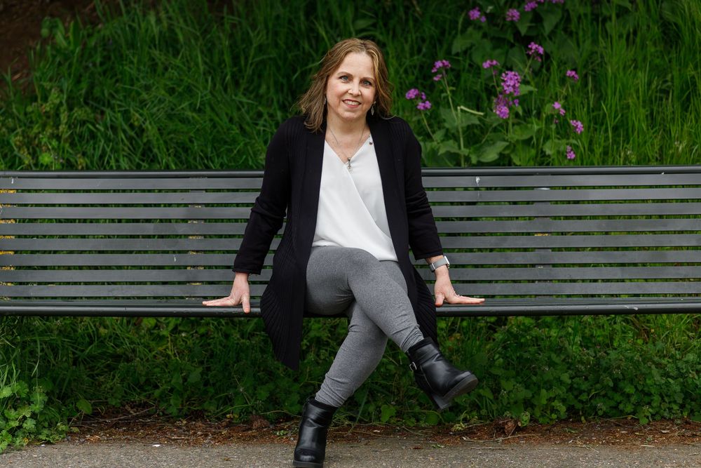 Woman in black blazer and gray pants sitting relaxed on a park bench surrounded by lush green foliage.