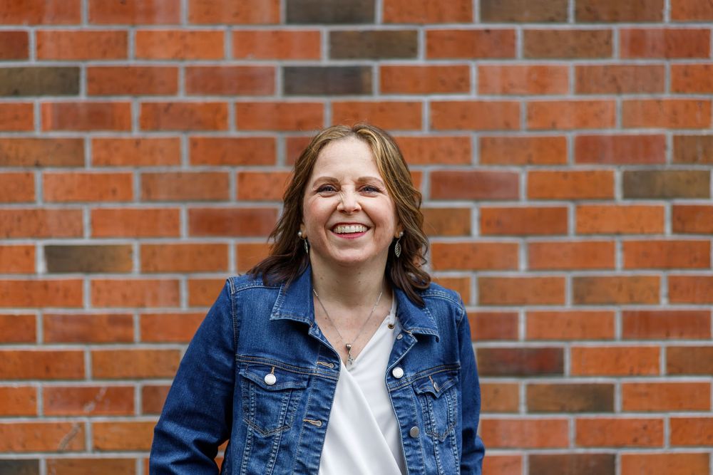 Smiling woman in denim jacket standing against a red brick wall outdoors.