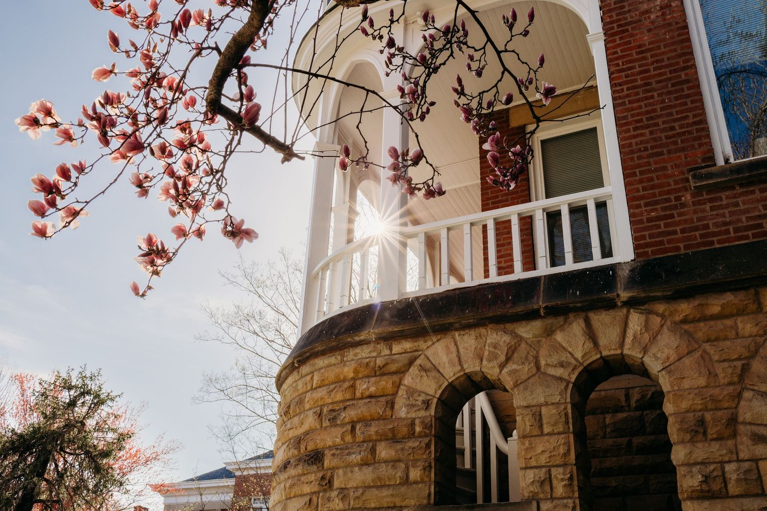 Red blooming spring flowers frame a vintage stone building with brick upper level and white balcony in sunlight. Dennison University