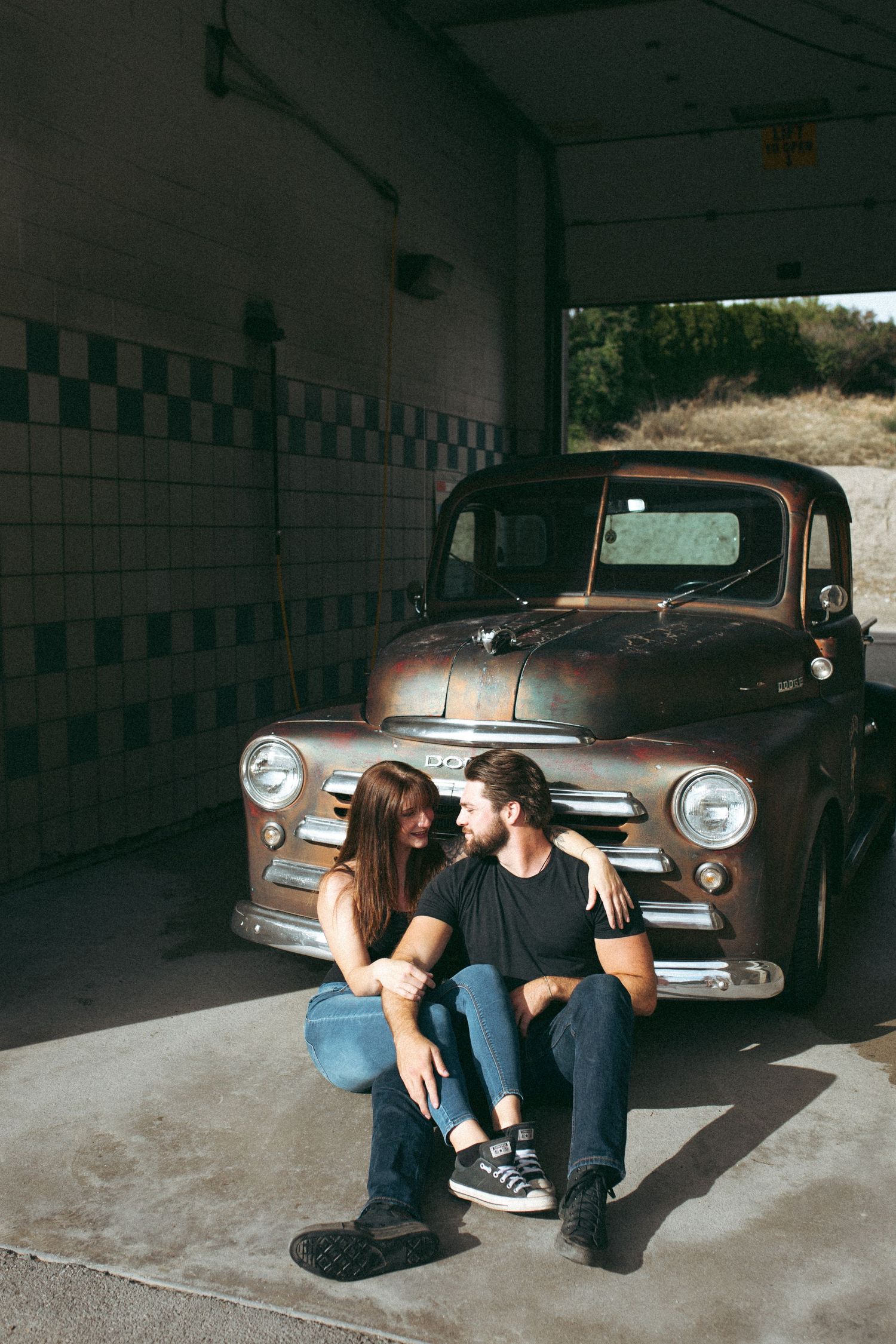 Couple sitting together in front of a rusty vintage pickup truck in a garage.