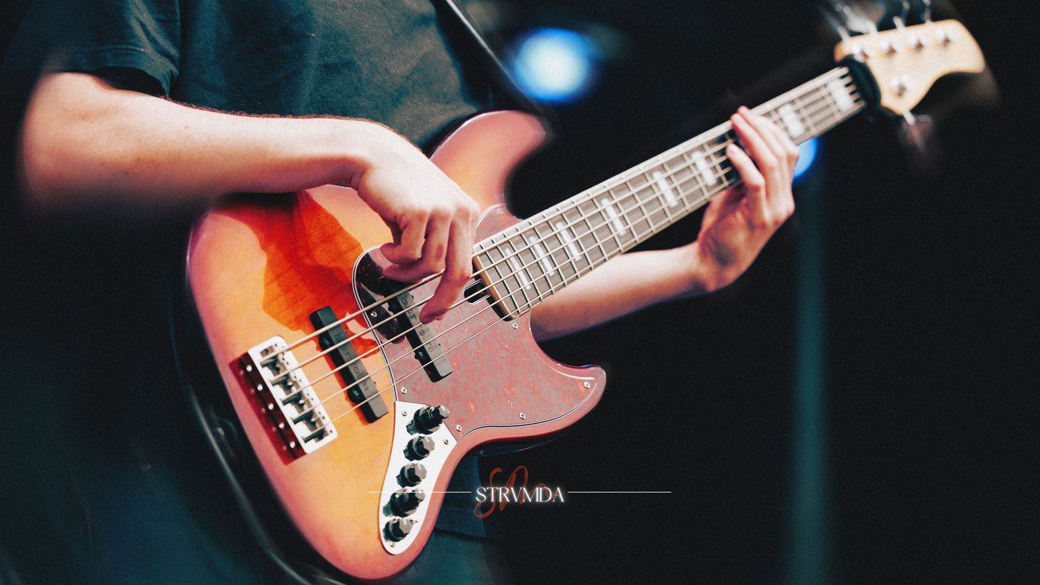 A musician plays an electric bass guitar on stage, with colorful sunburst finish under blue stage lighting.