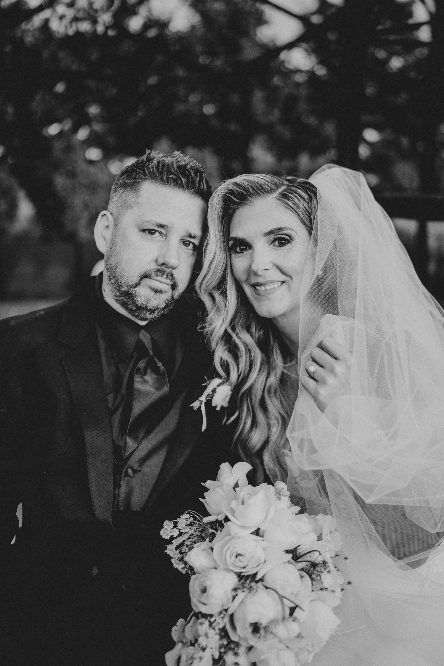 Black and white wedding portrait of a smiling bride in veil holding bouquet and groom in suit posing together outdoors.