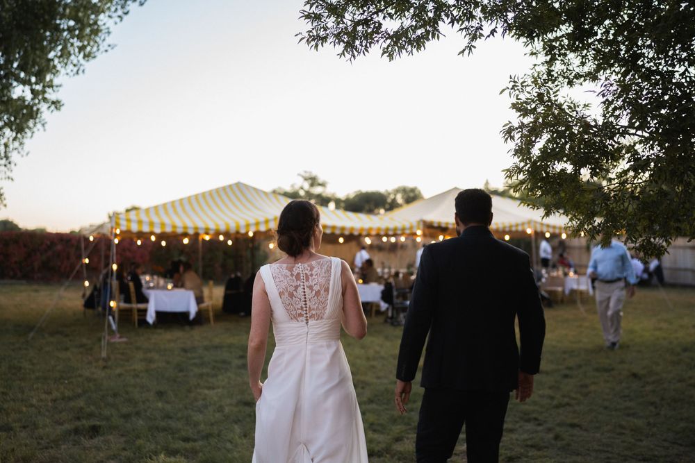 Bride and groom walking hand in hand toward a beautifully lit wedding reception tent at dusk at a backyard elopement