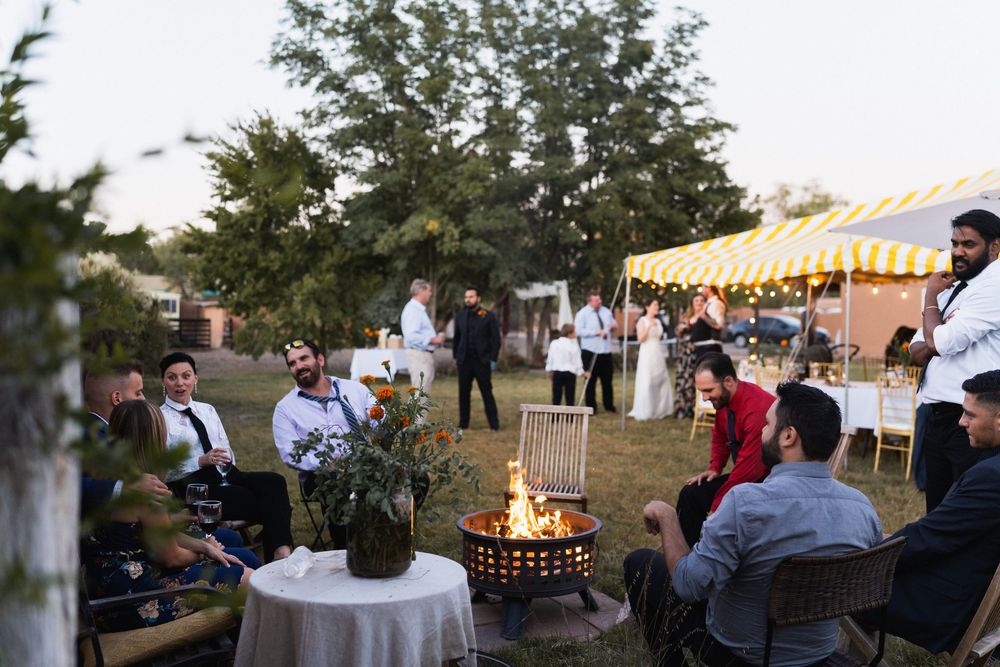 Wedding guests relax around a glowing fire pit at dusk with a yellow striped tent and string lights behind at a backyard elopement