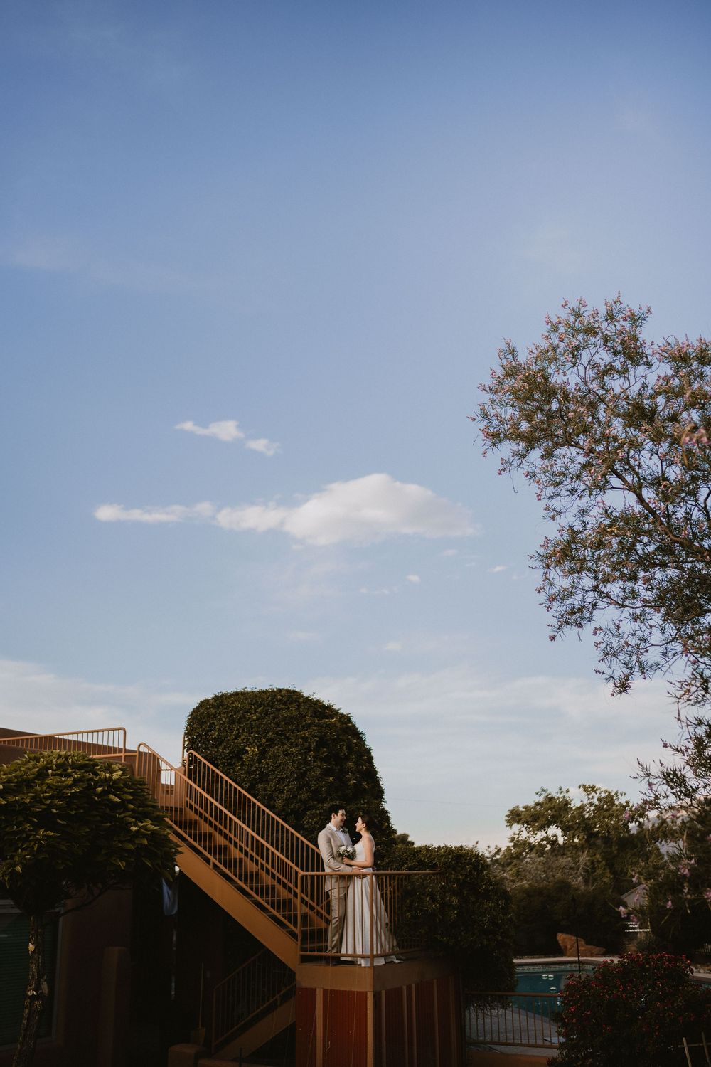 Bride and groom embracing on a staircase against a bright blue sky at golden hour, backyard elopement