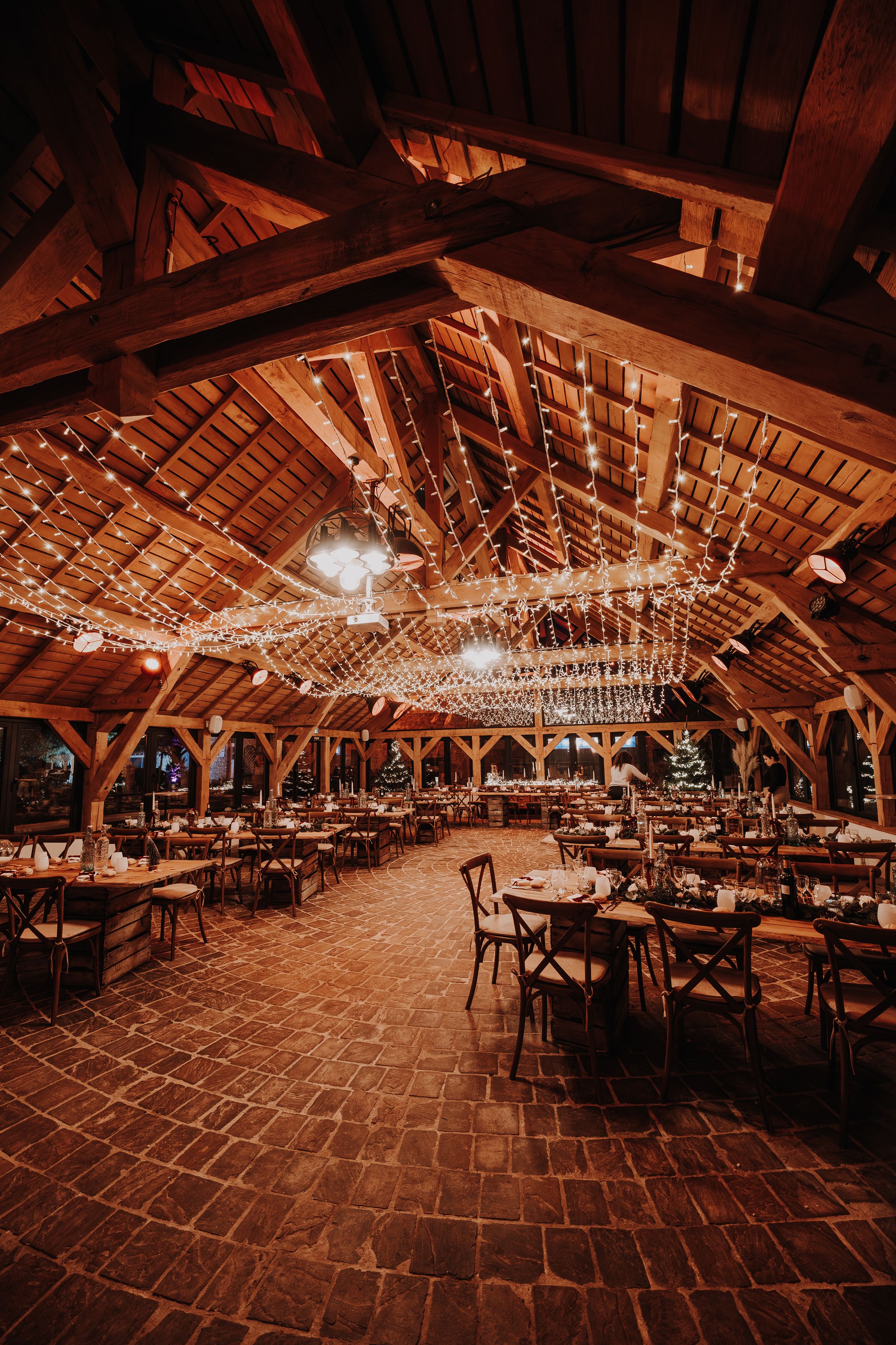 Rustic barn wedding venue with string lights, wooden beamed ceiling, brick floors, and elegantly set dining tables.