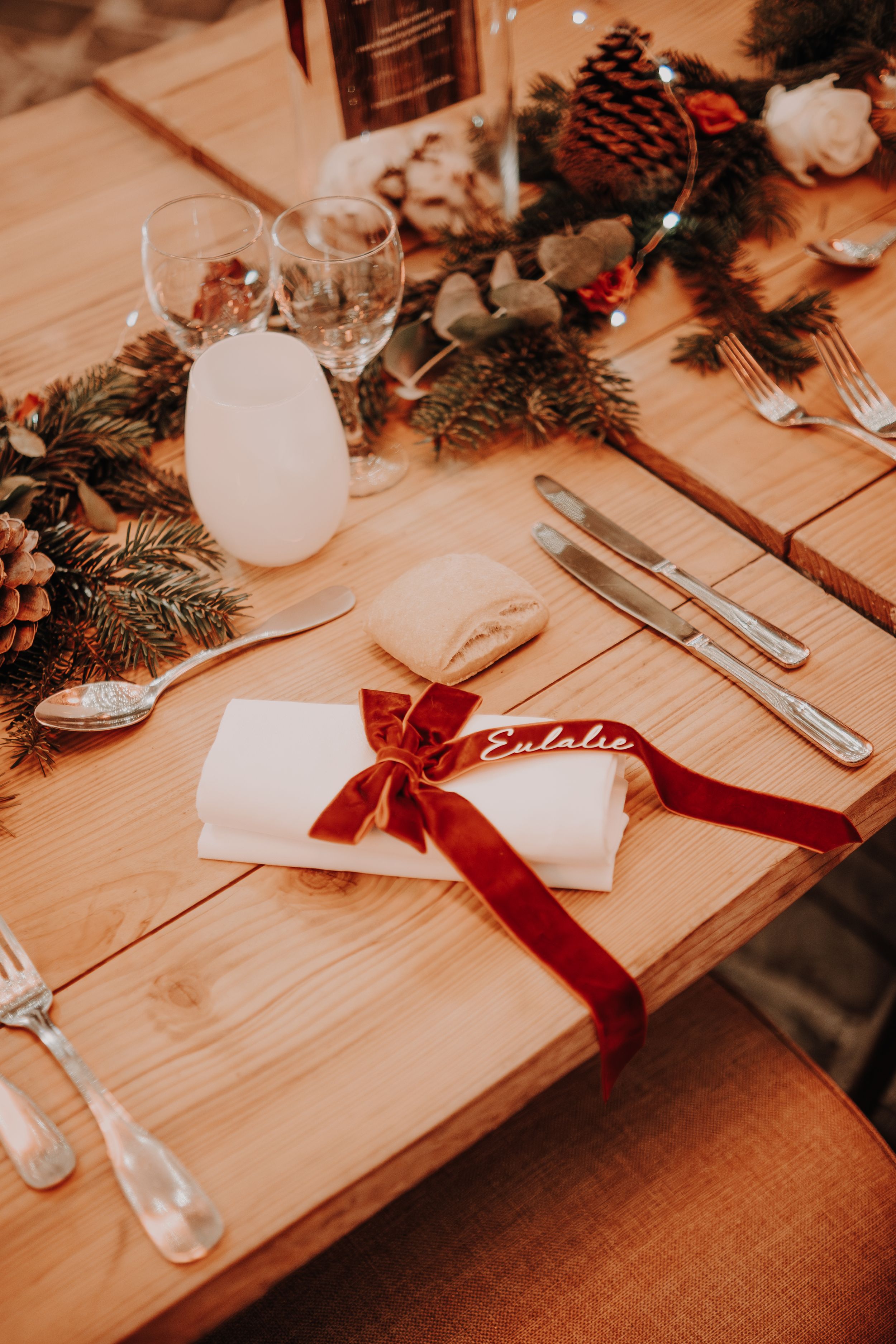 Elegant Christmas wedding table setting with pine greenery, red velvet ribbon name card, and silverware on rustic wood.