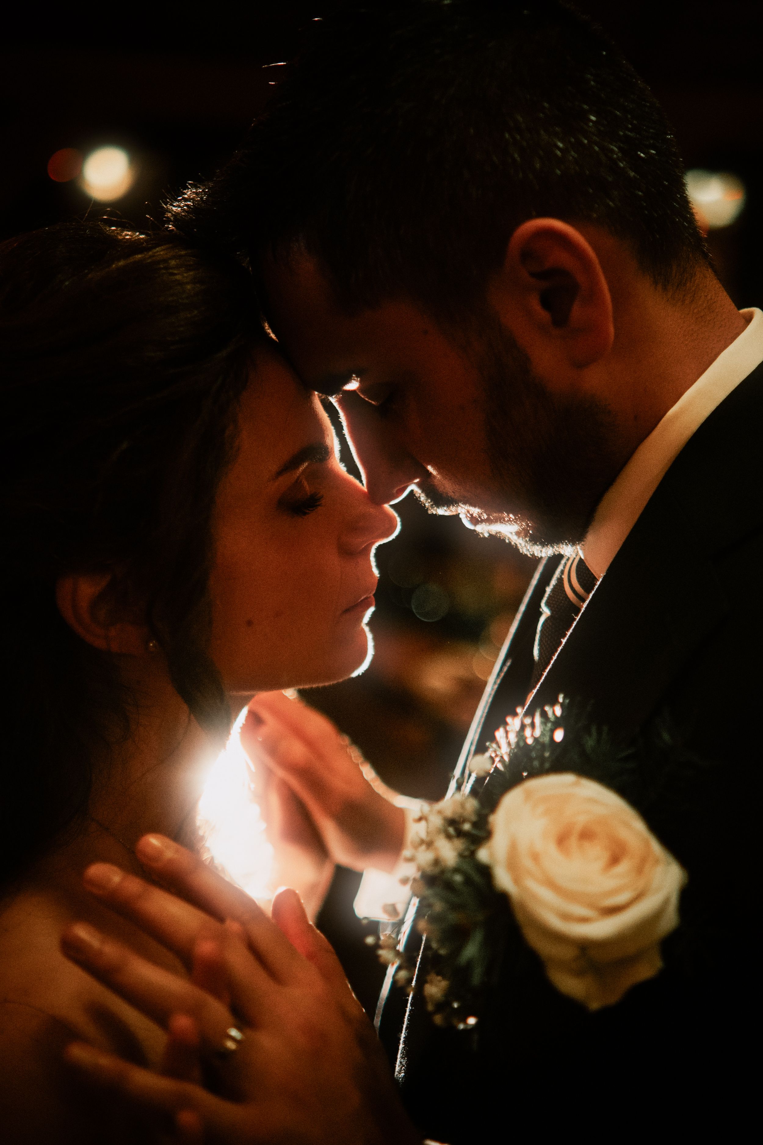 Bride and groom share an intimate moment, foreheads touching, illuminated by soft candlelight with a white rose boutonniere.