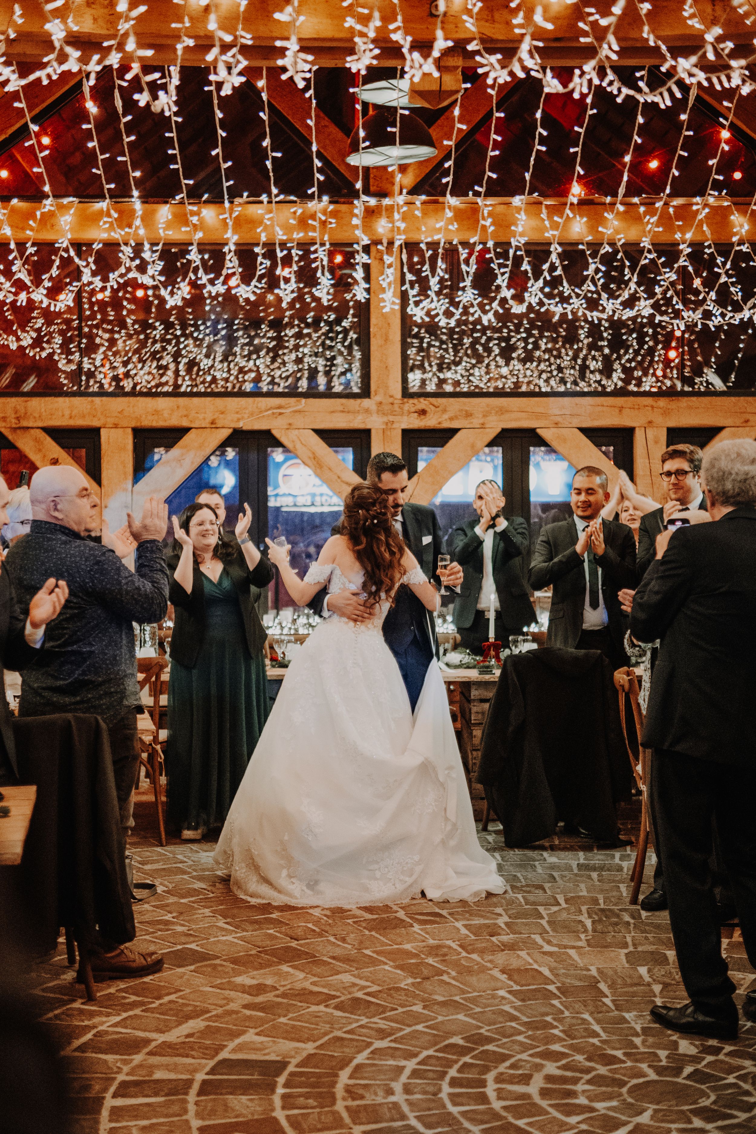 Bride and groom share first dance in a rustic barn venue adorned with twinkling fairy lights while guests applaud.