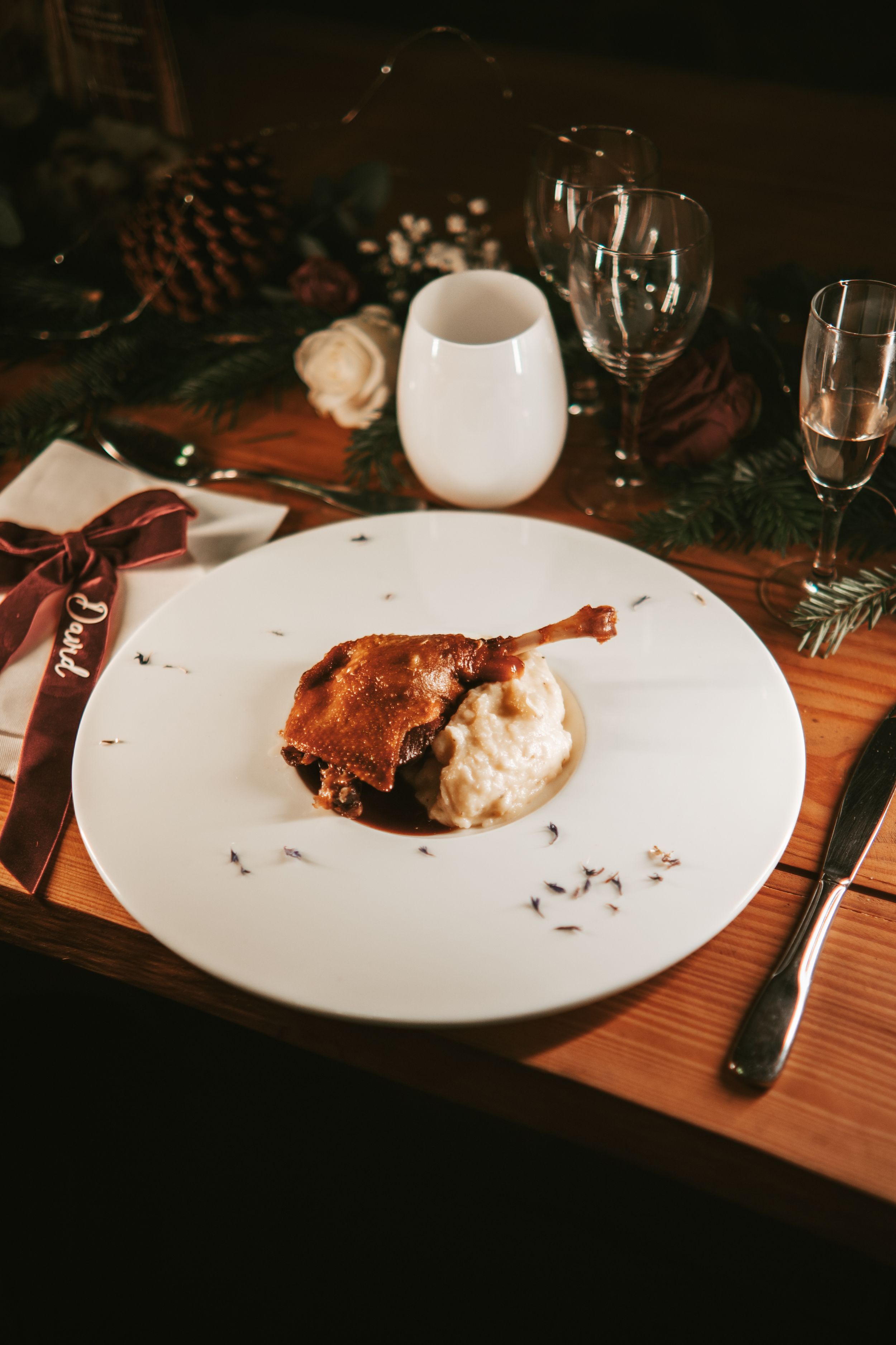 Elegant holiday dinner plate with roasted duck leg on creamy mash, set on a festive wooden table with wine glasses.