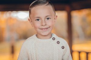 Smiling young boy with short hair wearing a cream cable-knit sweater in an outdoor autumn setting.